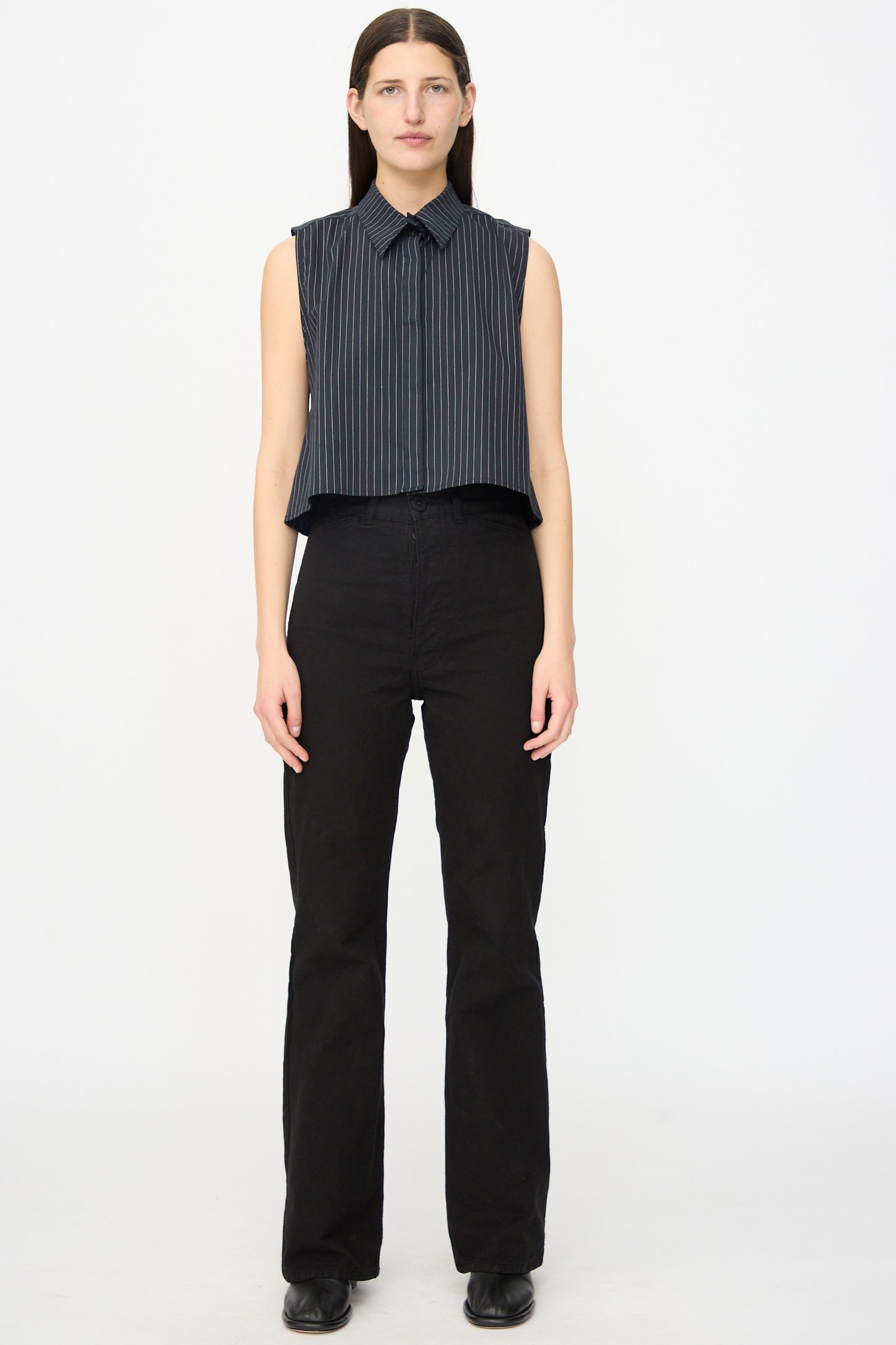 A person faces forward in a sleeveless, dark pinstriped shirt and Jesse Kamm’s Canvas Les Deux Pants in Black, paired with black shoes, standing against a plain white background.