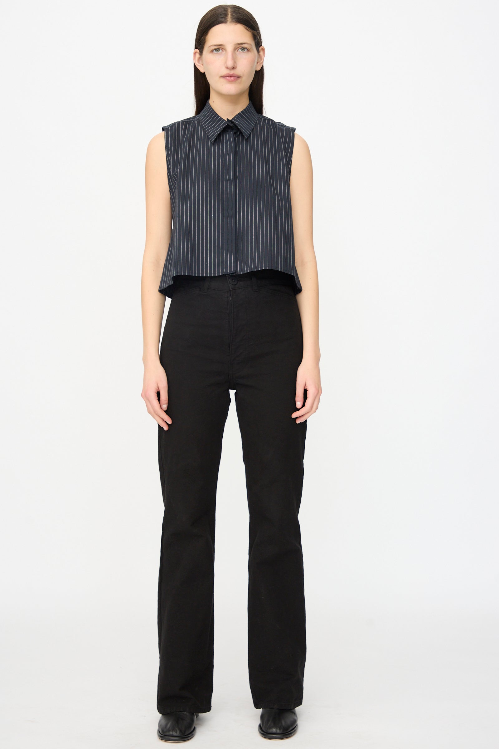 A person faces forward in a sleeveless, dark pinstriped shirt and Jesse Kamm’s Canvas Les Deux Pants in Black, paired with black shoes, standing against a plain white background.