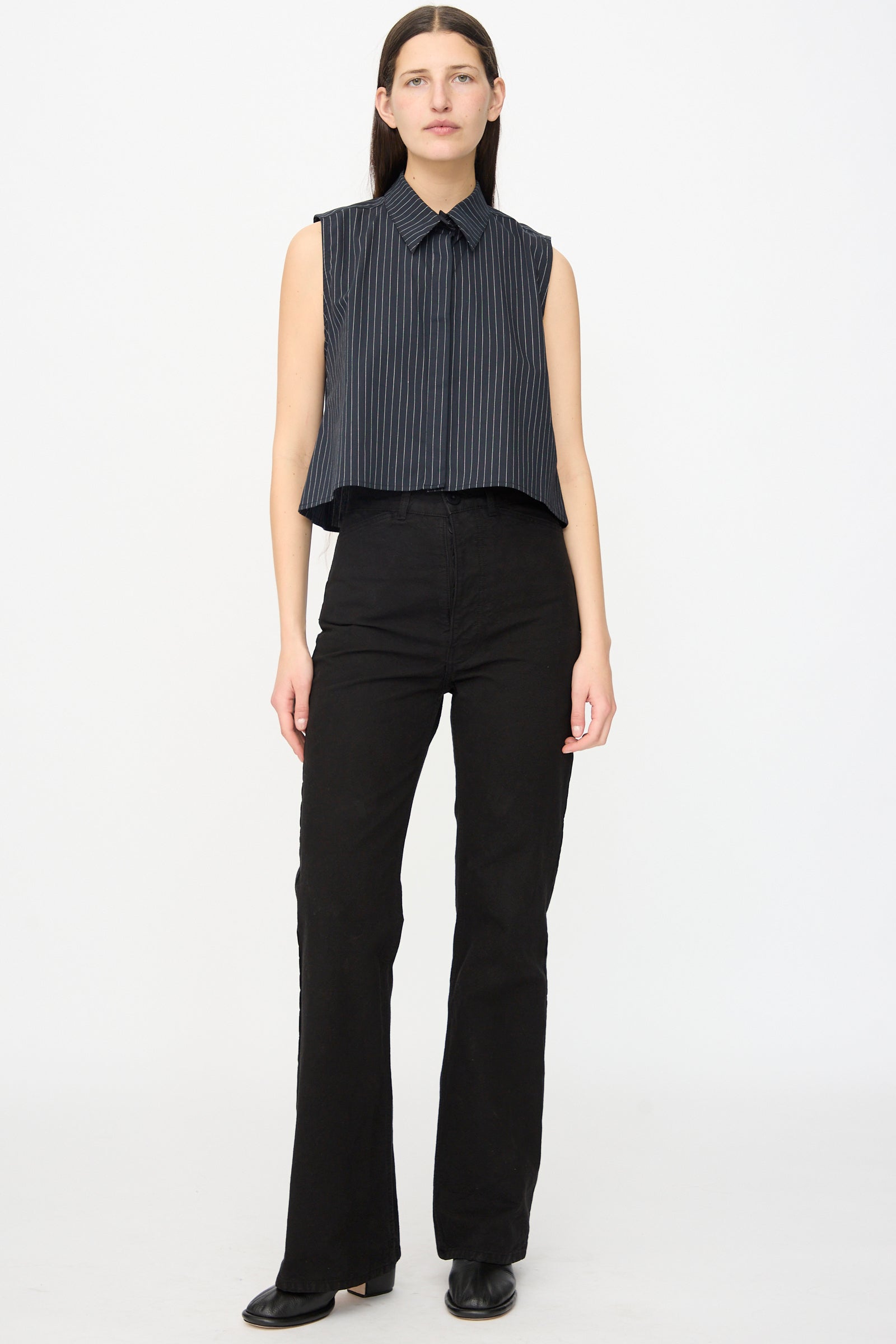 A person stands facing forward in a sleeveless, dark pinstripe collared shirt and Jesse Kamm Canvas Les Deux Pants in Black, paired with black shoes, against a plain white background.