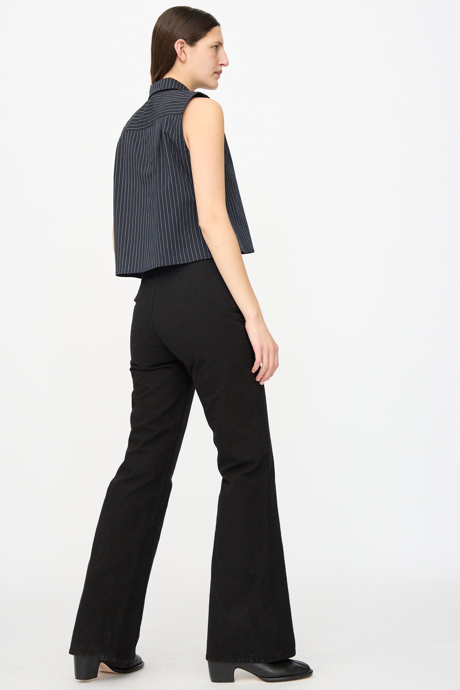A person stands sideways against a white background, wearing a sleeveless navy pinstripe top and Jesse Kamm Canvas Les Deux Pants in Black with black shoes.