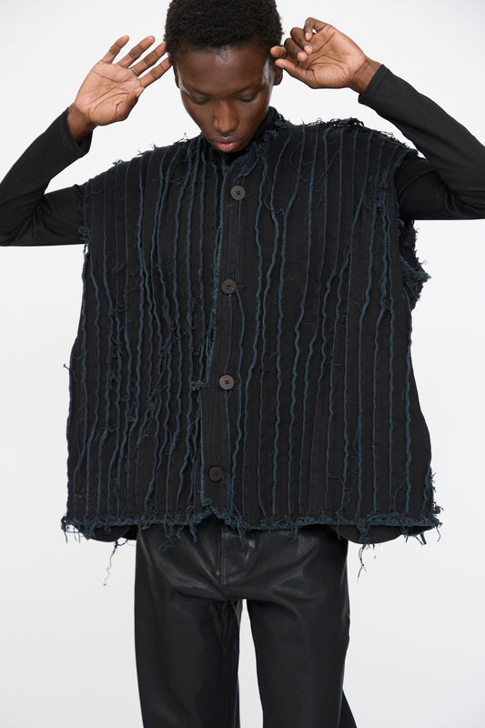 A person wearing the Jesse Kamm Deadstock 10oz. Denim Baleen Vest in Black—deconstructed, oversized, with vertical frayed lines—layered over a long-sleeve black shirt and black pants, stands against a plain background.