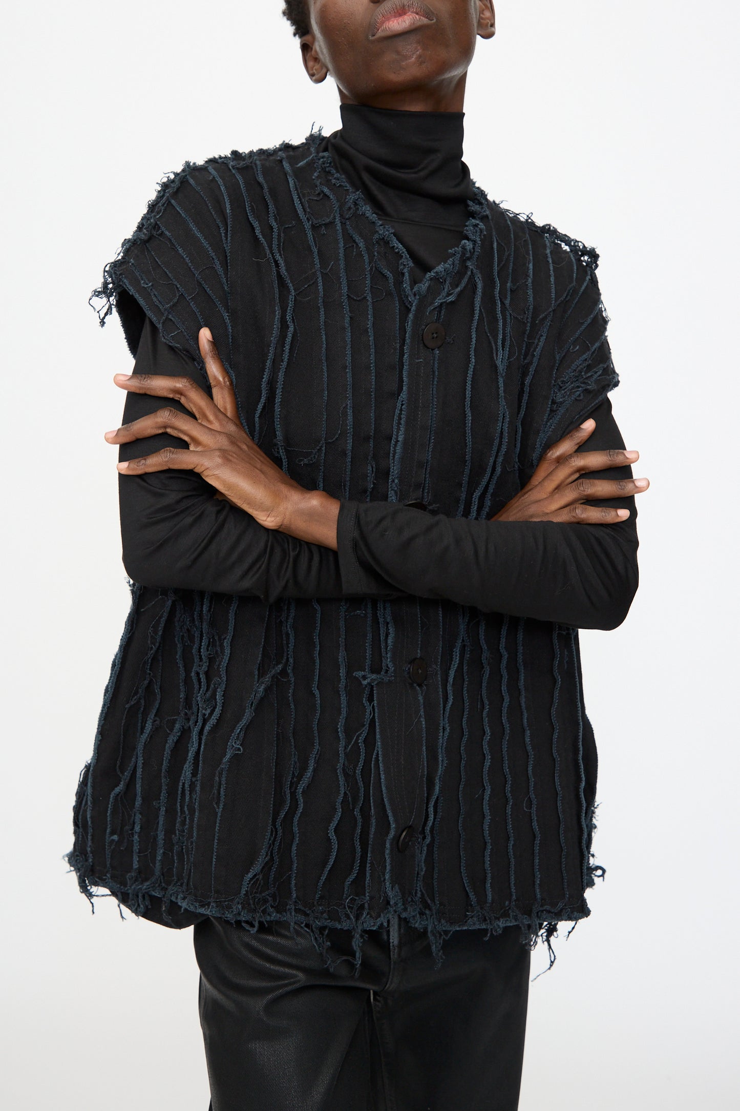 Model wears the Deadstock 10oz. Denim Baleen Vest in Black by Jesse Kamm—a textured, sleeveless, deconstructed style with frayed vertical lines—layered over a black turtleneck and black pants, arms crossed against a plain background.