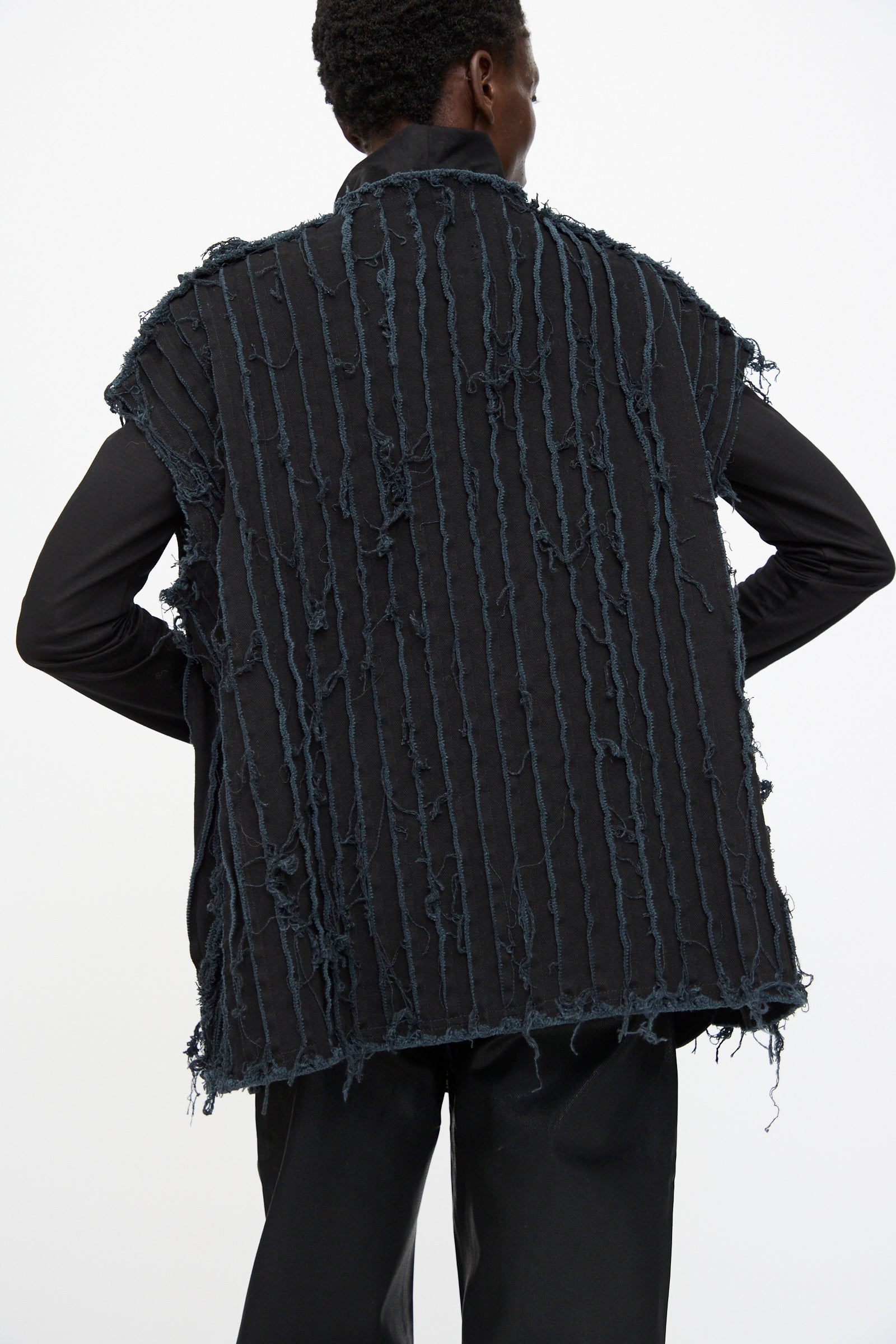 A person is seen from behind wearing the Jesse Kamm Deadstock 10oz. Denim Baleen Vest in Black over a long-sleeve black shirt and black pants, featuring vertical frayed textured lines.