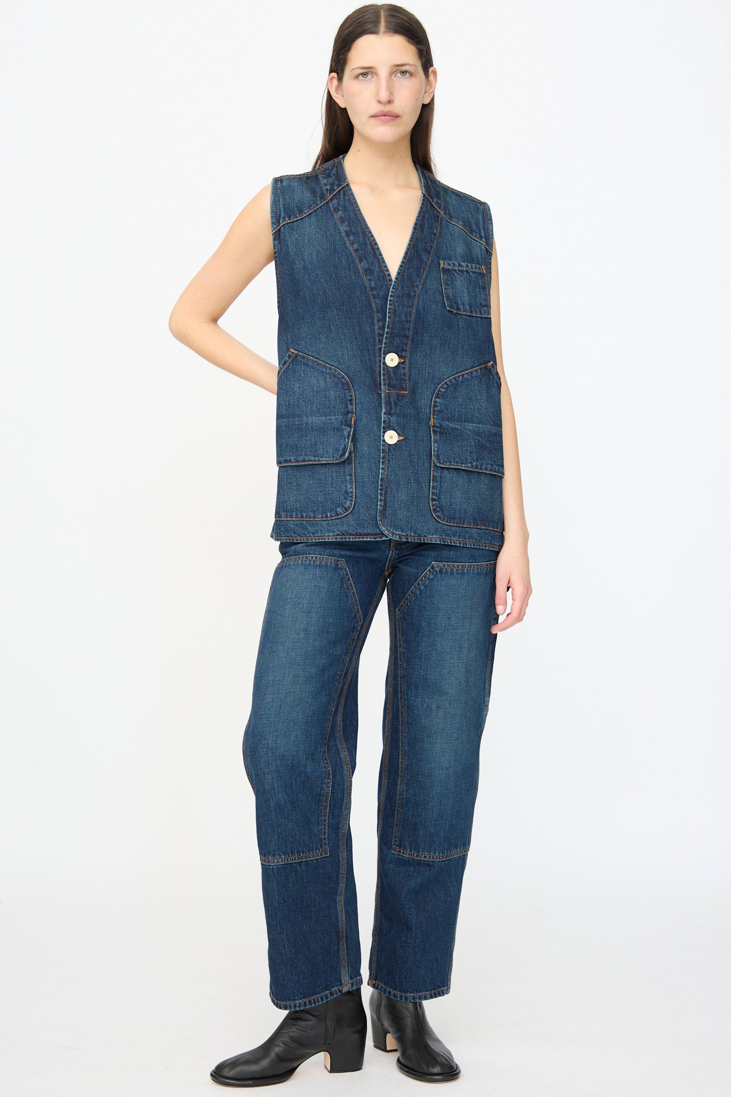 A person stands against a plain background wearing the Jesse Kamm Denim Directors Vest in Medium Roast, featuring large pockets, paired with matching denim pants and black boots.