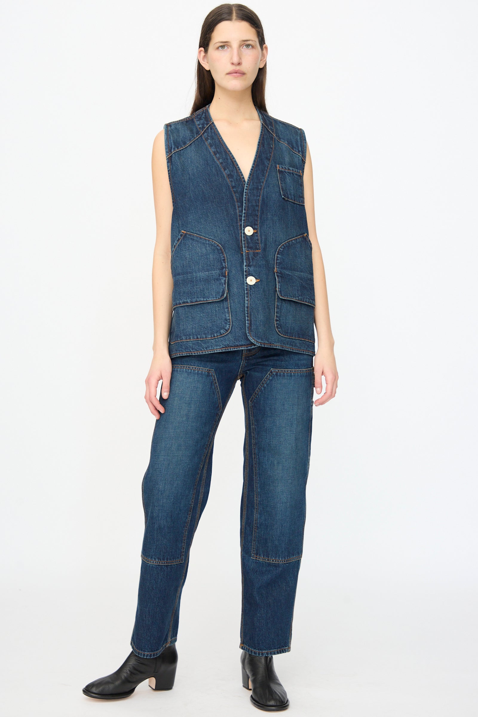 A person wears the Jesse Kamm Denim Directors Vest in Medium Roast— a sleeveless, button-up piece with large pockets — along with matching denim pants and black boots, standing against a plain white background.