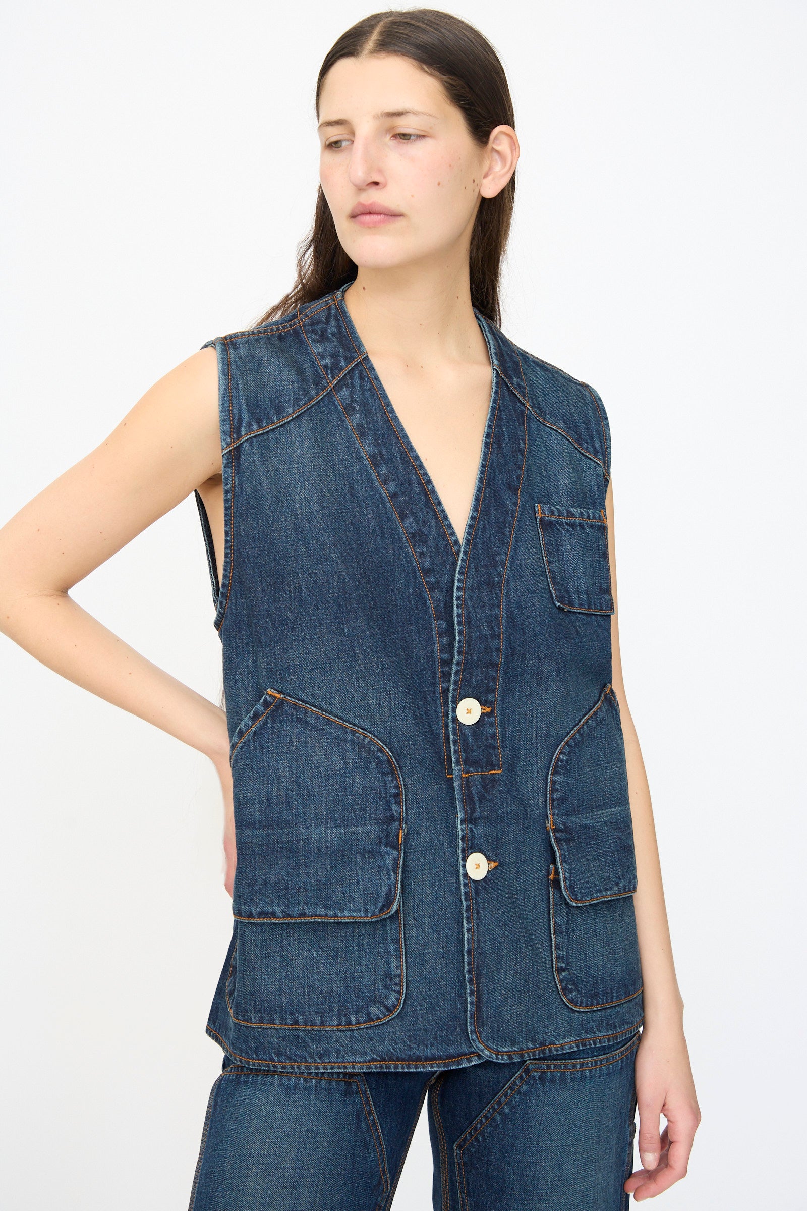 A woman wears the Jesse Kamm Denim Directors Vest in Medium Roast—sleeveless, button-up, with front pockets—paired with matching denim pants, standing against a plain white background.