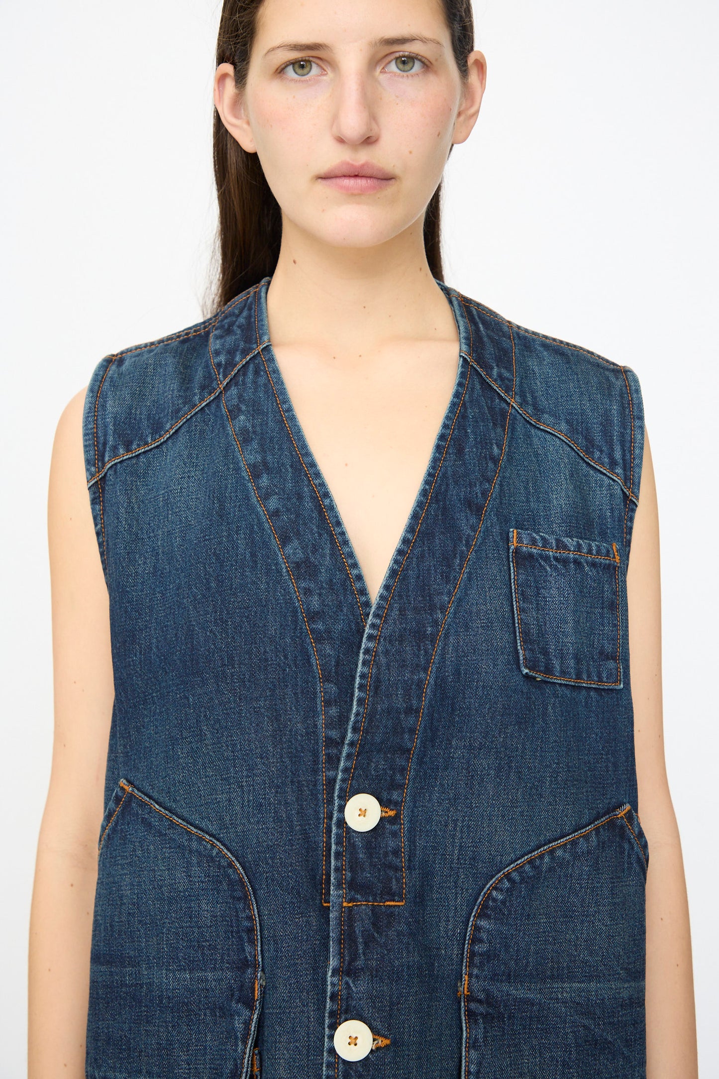 A person wearing the Jesse Kamm Denim Directors Vest in Medium Roast, a sleeveless, button-up piece made from regenerative Japanese denim with large front pockets and white buttons, stands against a plain background.