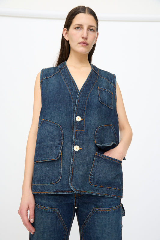 A woman wears the Jesse Kamm Denim Directors Vest in Medium Roast—sleeveless with large front pockets, crafted from regenerative Japanese denim—paired with matching pants, standing against a plain white background.