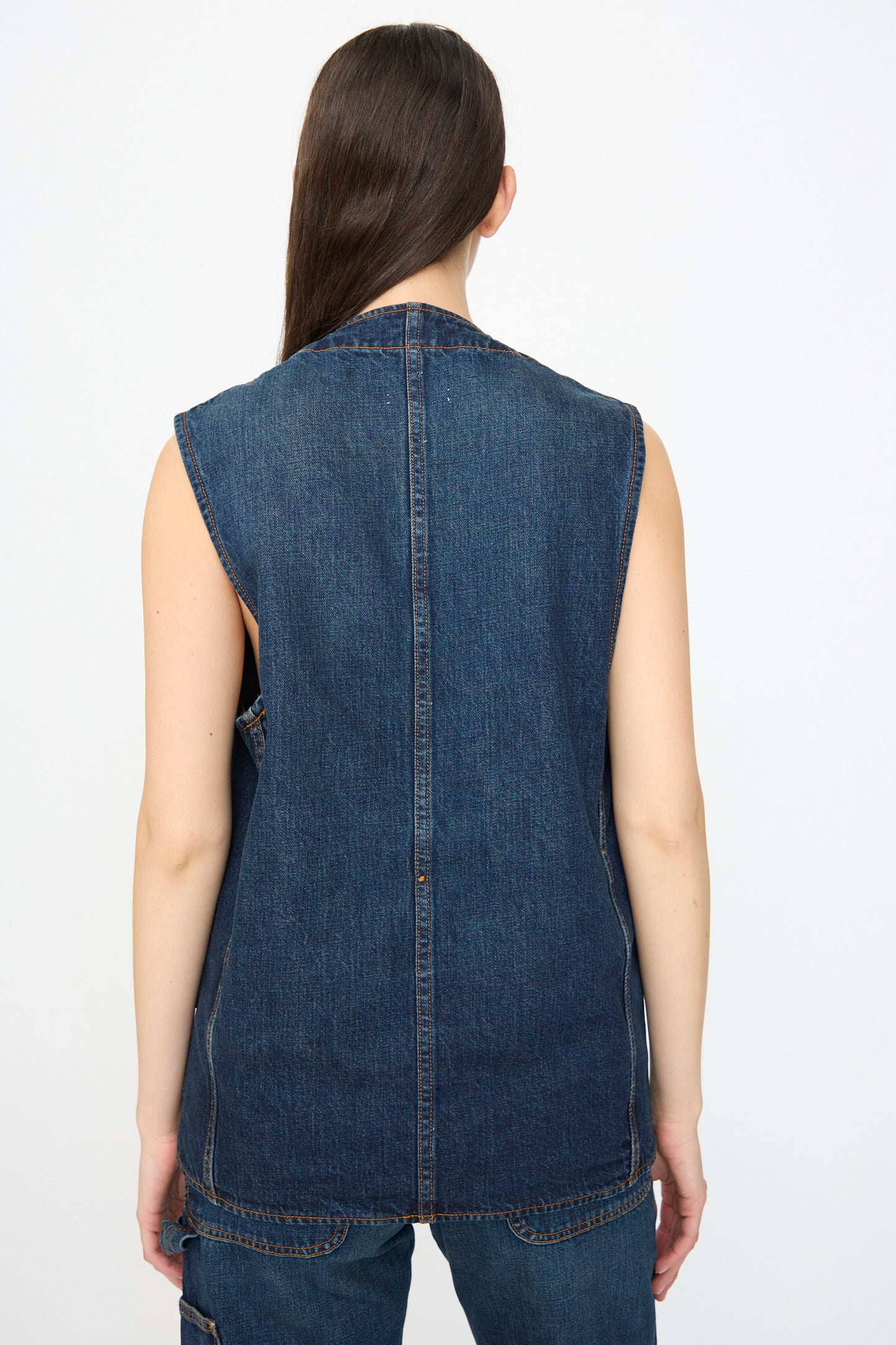 A person with long brown hair is shown from the back, wearing the Jesse Kamm Denim Directors Vest in Medium Roast and matching jeans made from regenerative Japanese denim, against a plain light background.