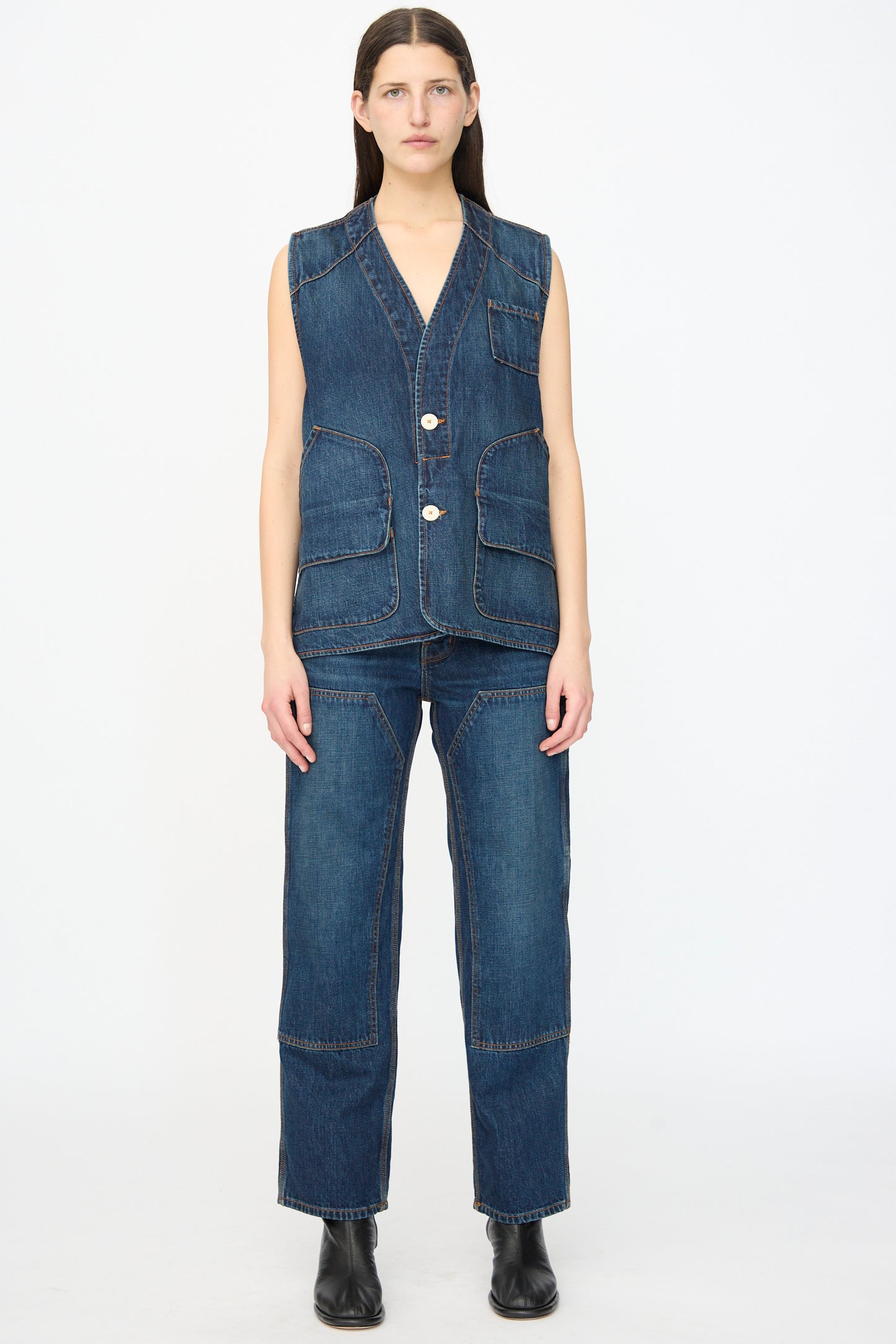 A person faces forward in Jesse Kamm's Denim Patchwork Handy Pants in Medium Roast, paired with a sleeveless denim vest and black boots against a plain white background.