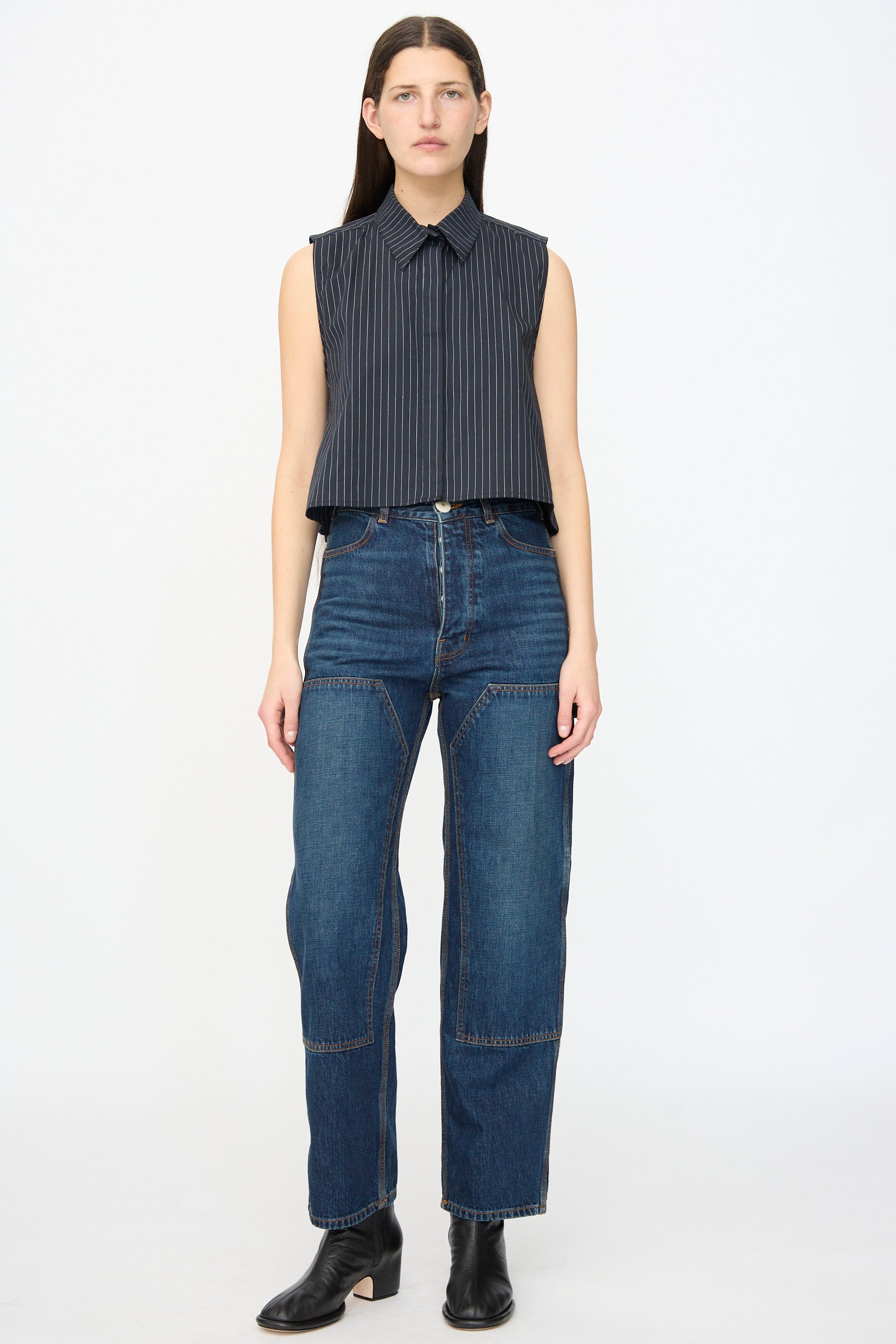 A person faces forward against a white background wearing Jesse Kamm Denim Patchwork Handy Pants in Medium Roast, styled with a sleeveless pinstripe collared top and black ankle boots.