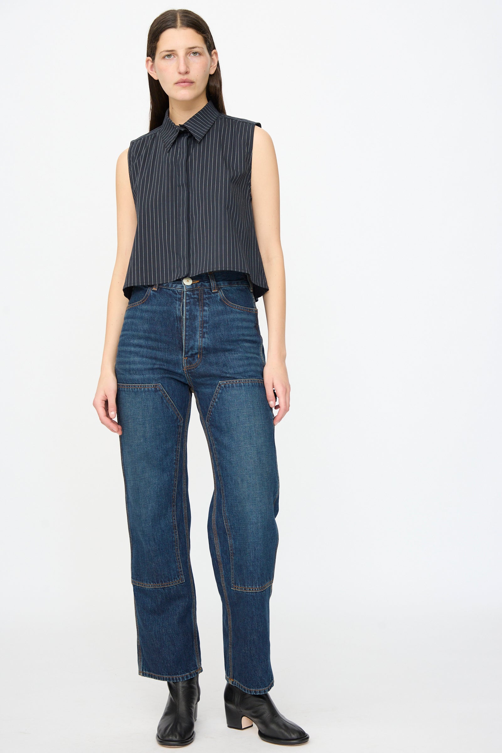 A woman stands against a plain background wearing a sleeveless pinstripe top, Jesse Kamm Denim Patchwork Handy Pants in Medium Roast, and black ankle boots.