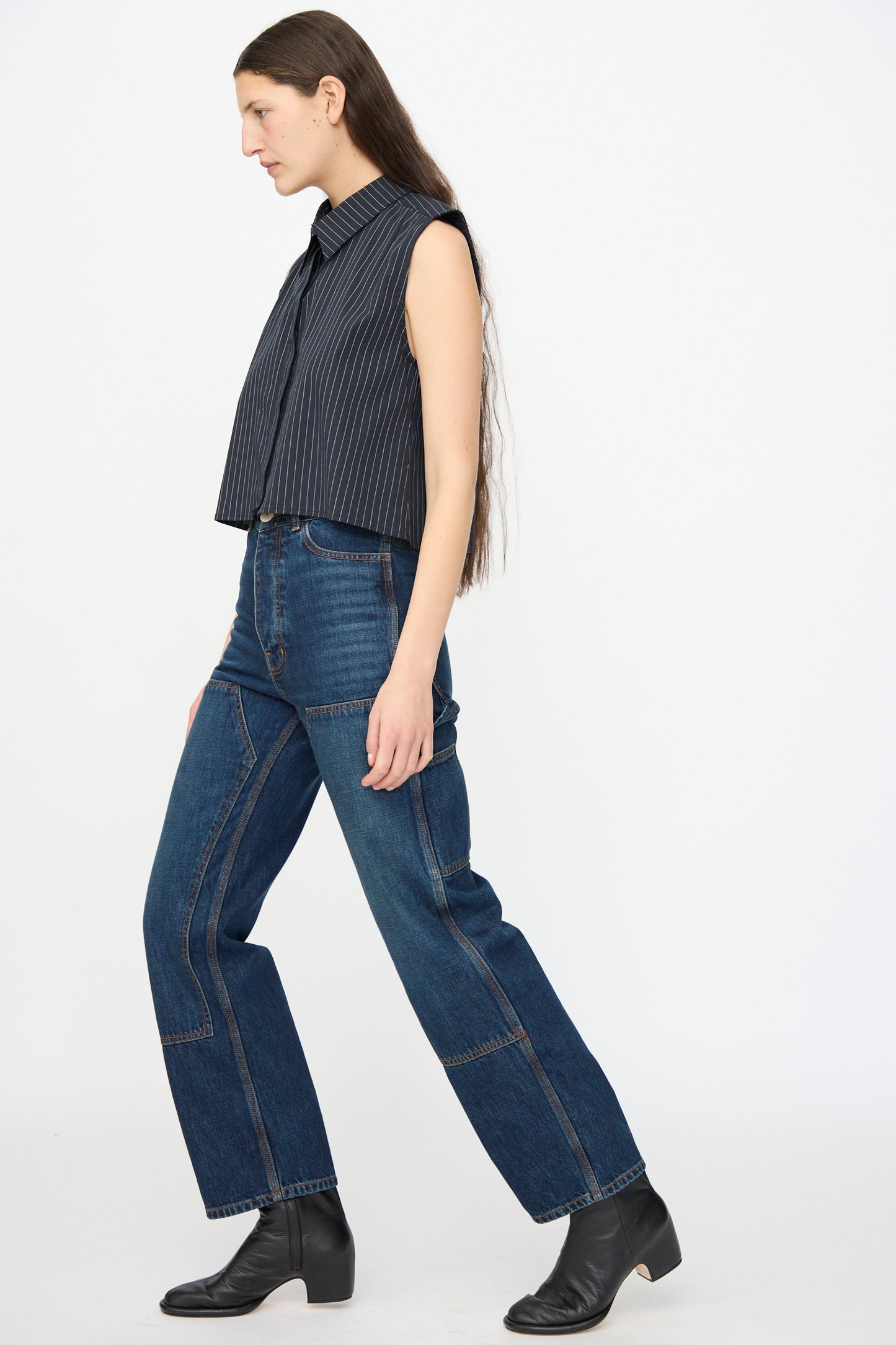A person with long hair wears a sleeveless pinstripe top, Jesse Kamm Denim Patchwork Handy Pants in Medium Roast, and black ankle boots, standing against a plain white background.