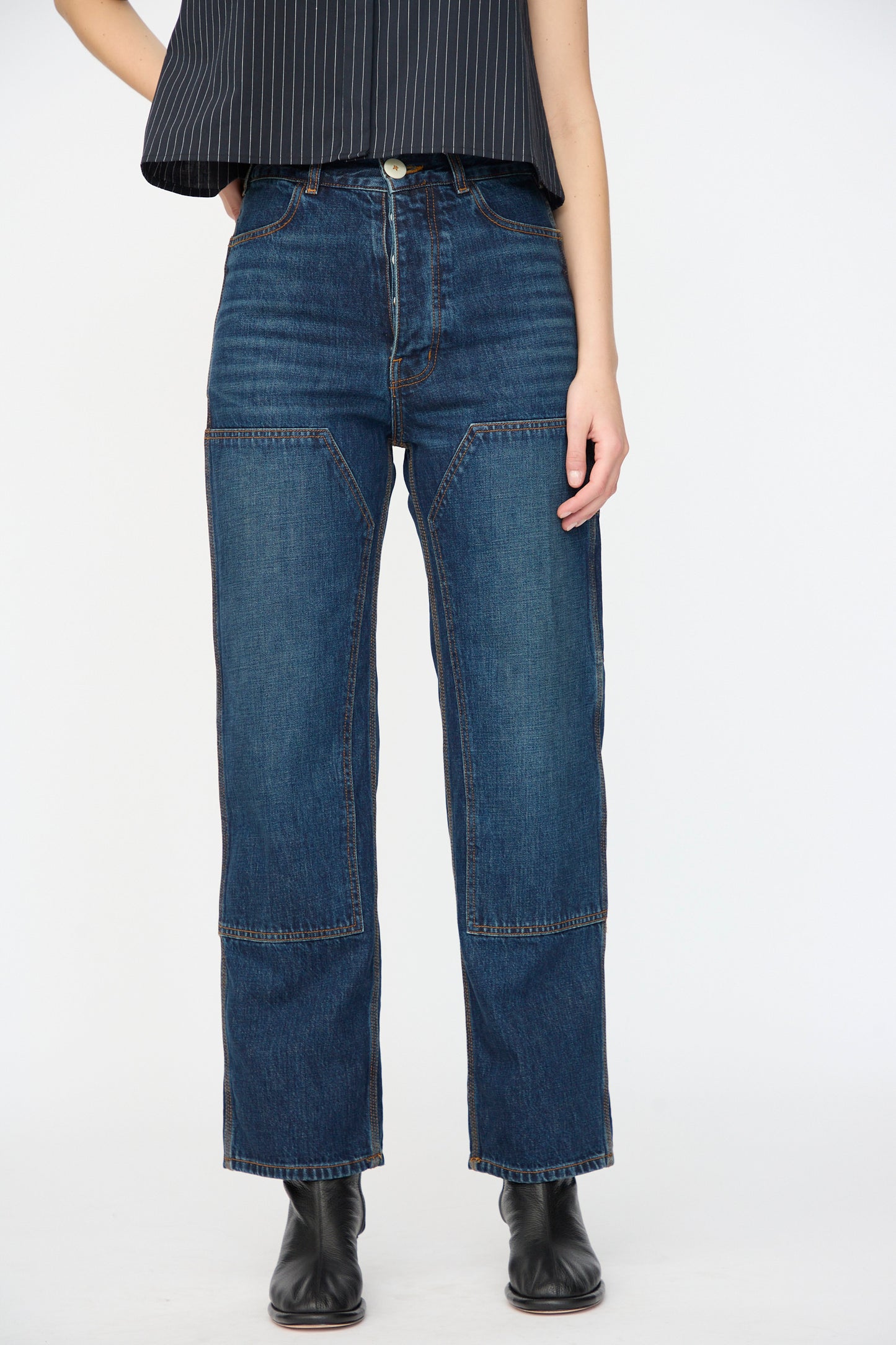 A person wears Jesse Kamm’s Denim Patchwork Handy Pants in Medium Roast with a black pinstriped top and black ankle boots, standing against a plain white background.