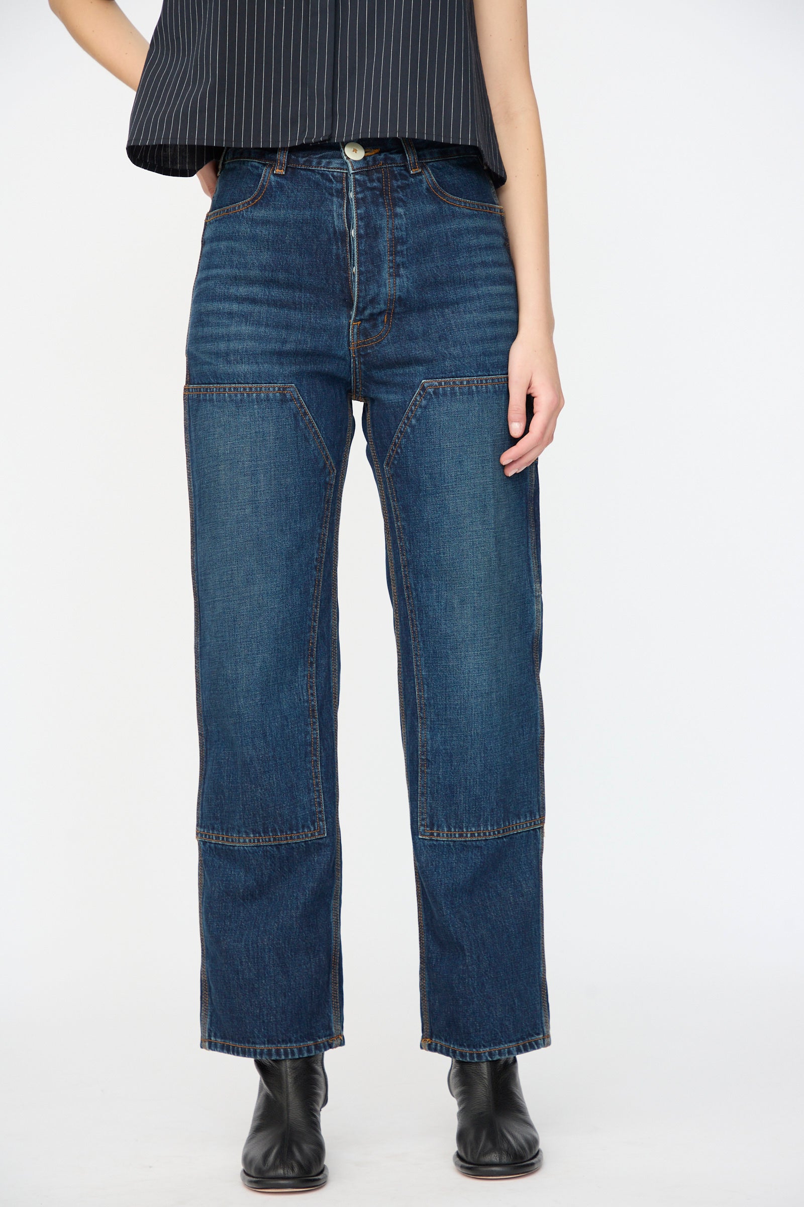 A person wears Jesse Kamm’s Denim Patchwork Handy Pants in Medium Roast with a black pinstriped top and black ankle boots, standing against a plain white background.