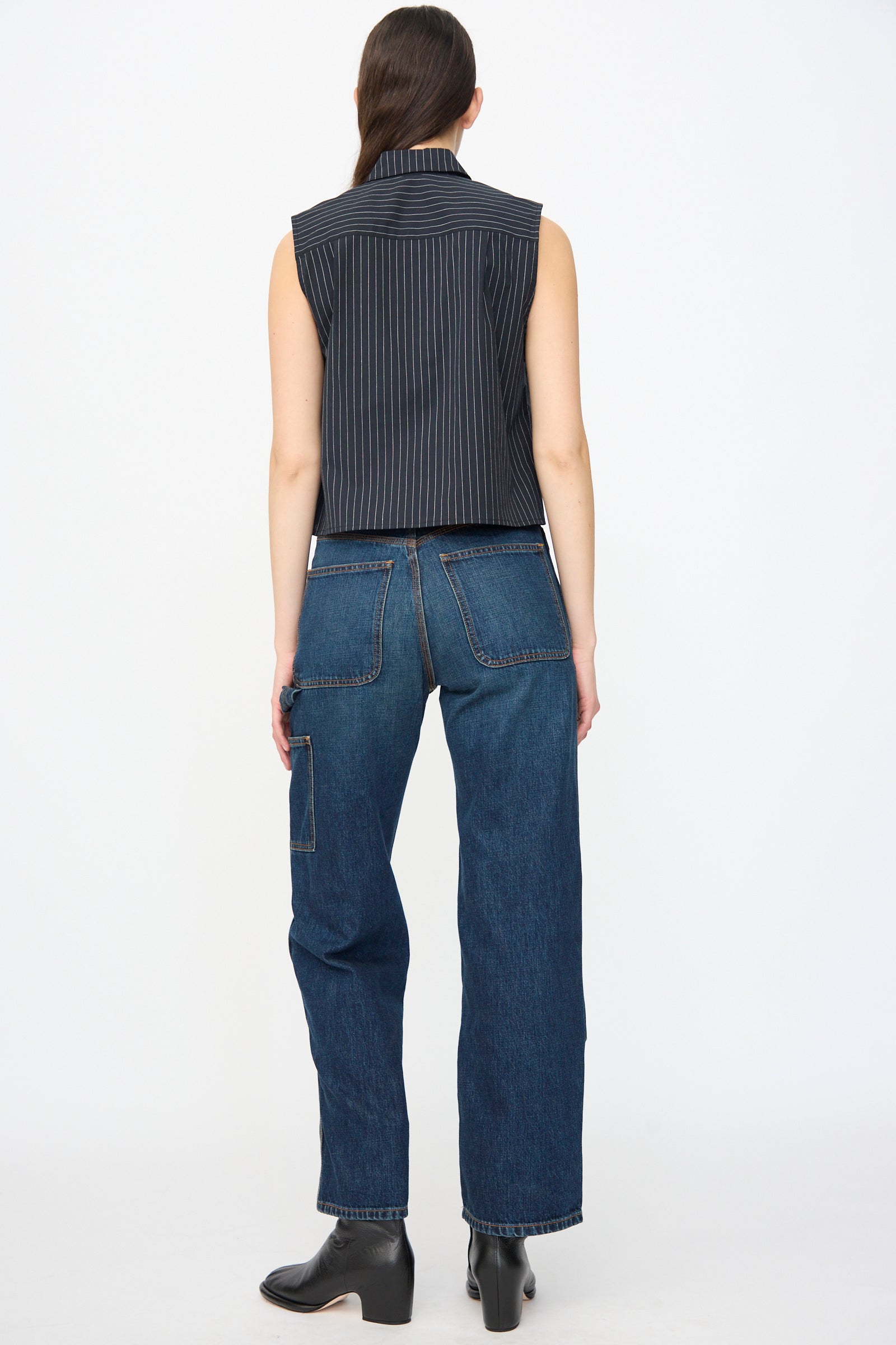 A person stands facing away, wearing a sleeveless pinstriped top, Jesse Kamm Denim Patchwork Handy Pants in Medium Roast with wide legs and large back pockets, paired with black ankle boots.