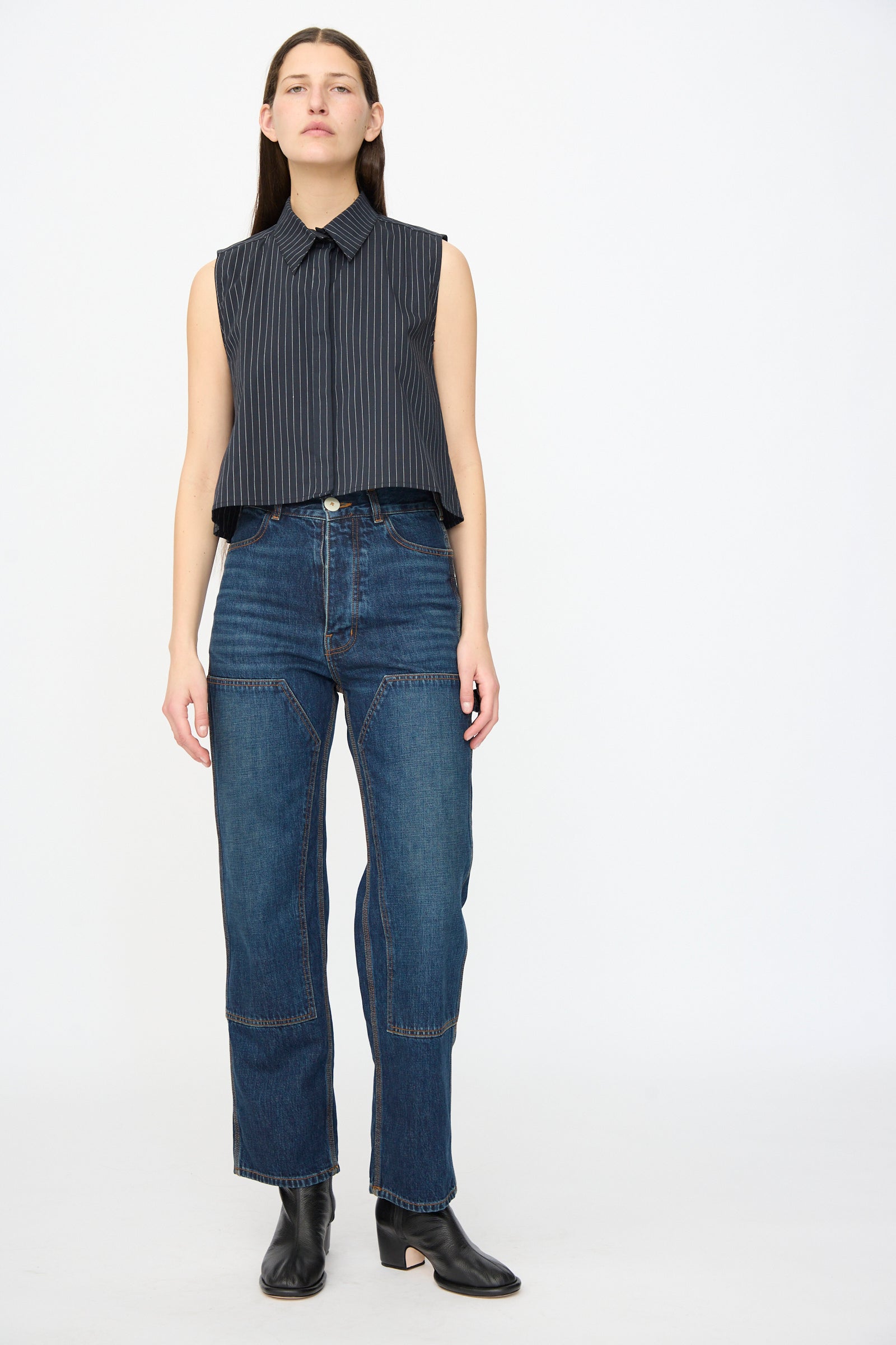 A woman stands against a plain background wearing the Jesse Kamm Ellis Top in Black with Fine Ivory Stripe, high-waisted patchwork jeans, and black ankle boots.