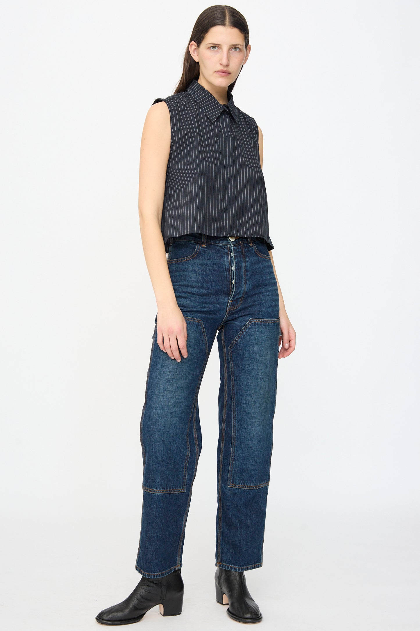 A person stands facing forward, wearing the Jesse Kamm Ellis Top in Black with Fine Ivory Stripe, high-waisted blue jeans with visible seams, black ankle boots, and has straight brown hair.
