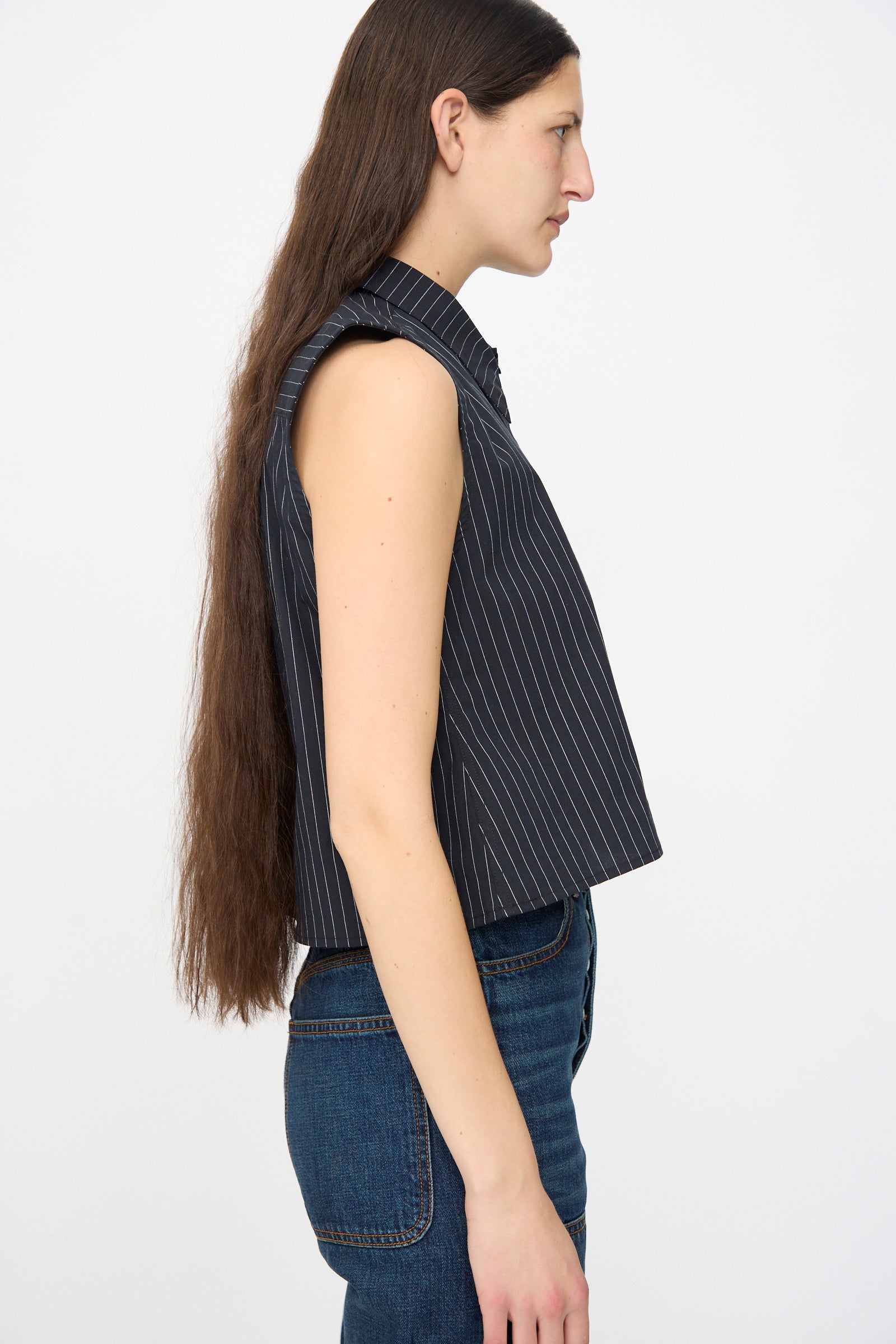 A person with long brown hair wears the Jesse Kamm Ellis Top in Black with Fine Ivory Stripe, styled in a cropped fit, along with blue jeans, standing in profile against a plain white background.