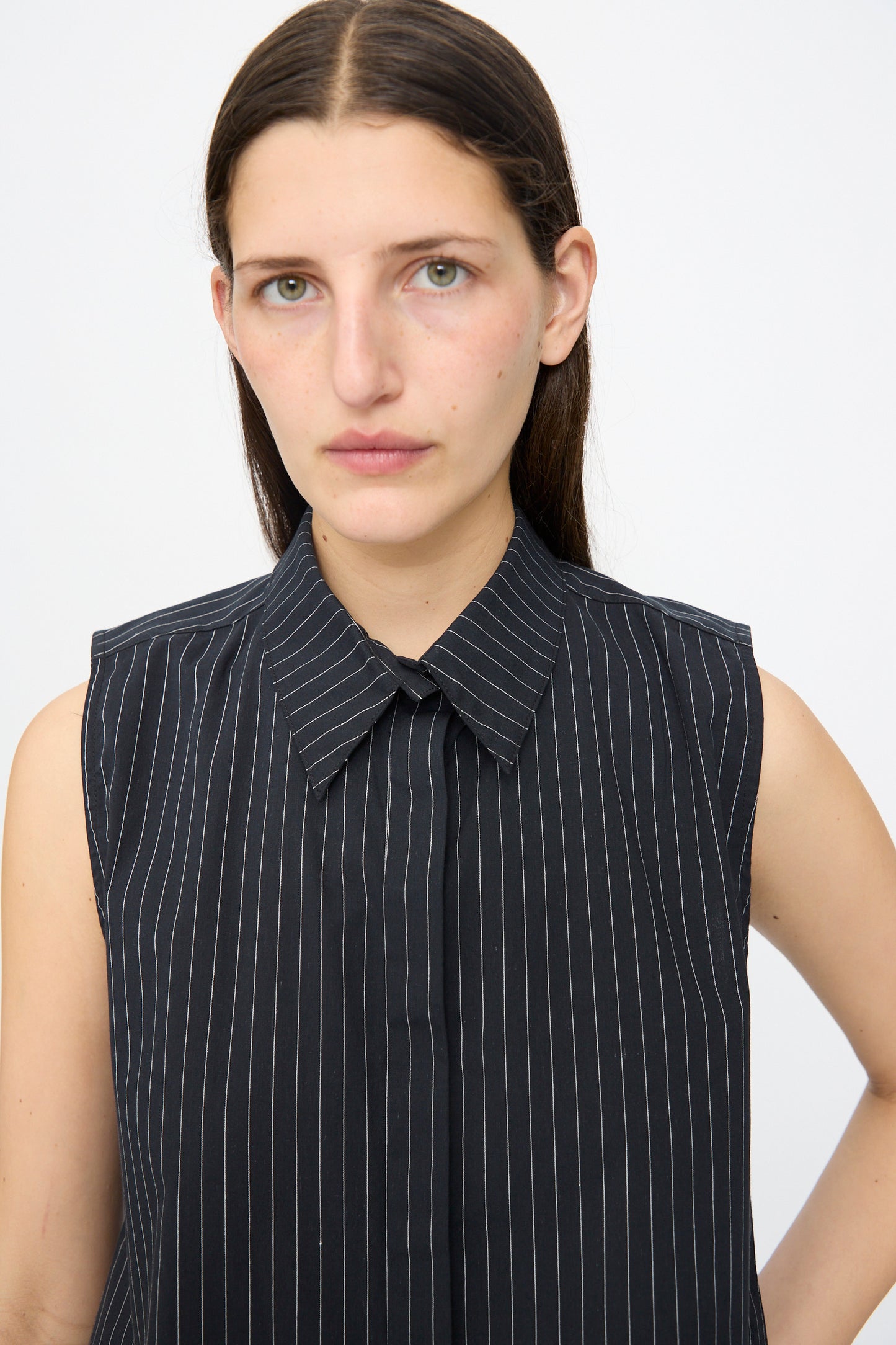 A person with straight brown hair and a neutral expression wears the Jesse Kamm Ellis Top in Black with Fine Ivory Stripe, featuring a sleeveless, cropped fit and pointed collar, standing against a plain light background.