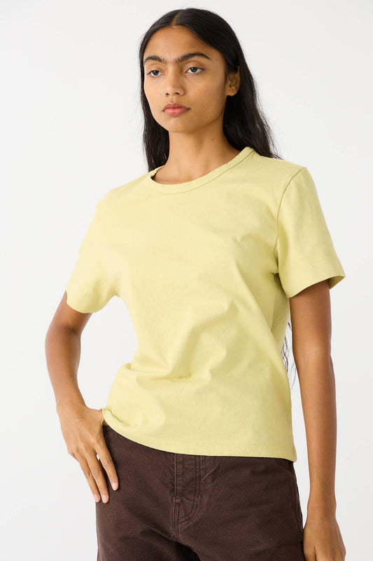 A person with long dark hair wearing a Jesse Kamm Fine Cotton Knit Sailor Tee in Citroen, featuring a light yellow hue and relaxed fit along with dark brown pants, stands with one hand on the hip and the other arm relaxed.