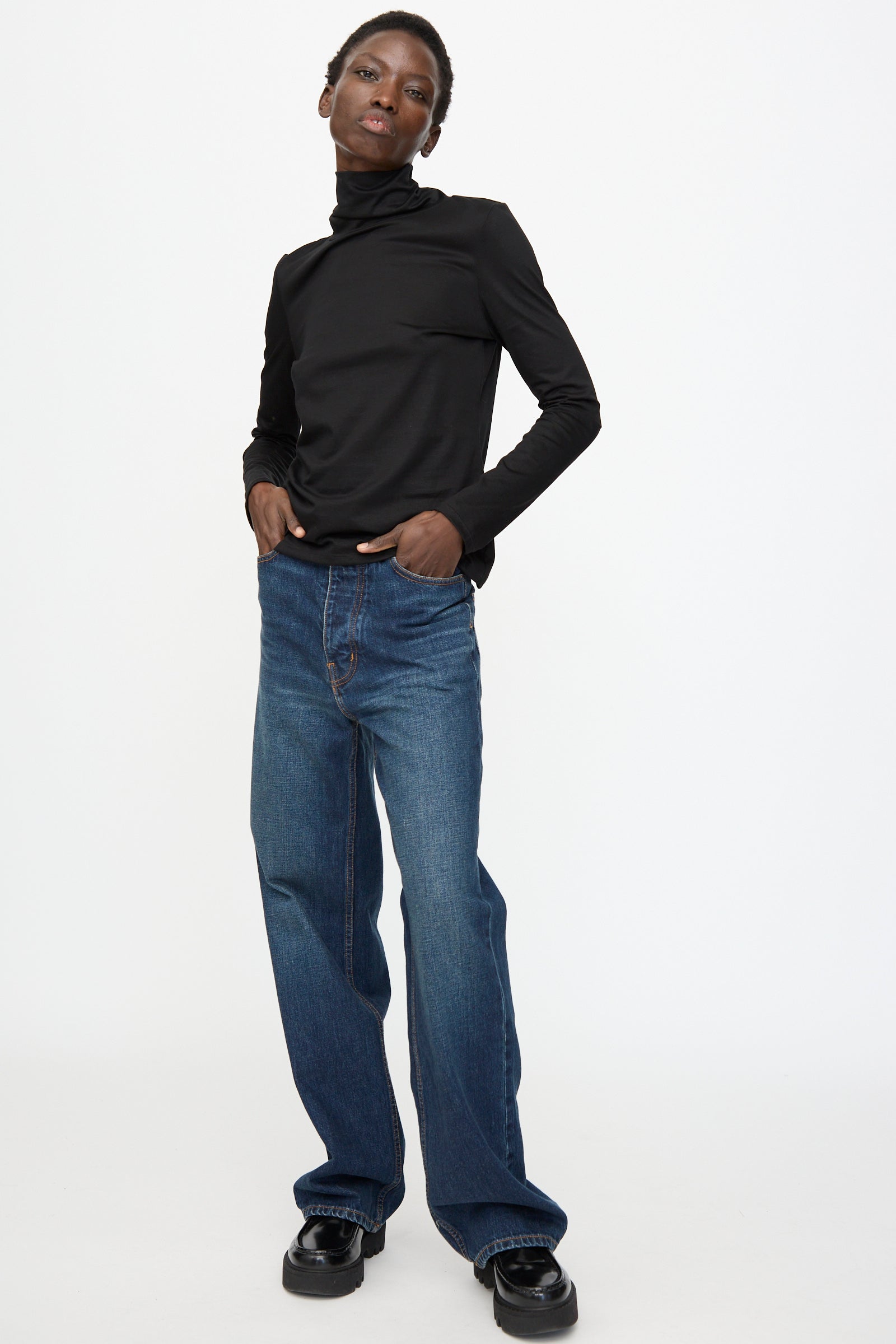 A person stands against a white background wearing a black turtleneck, black shoes, and Jesse Kamm Japanese Denim Alta Jeans in Medium Roast with one hand in their pocket.