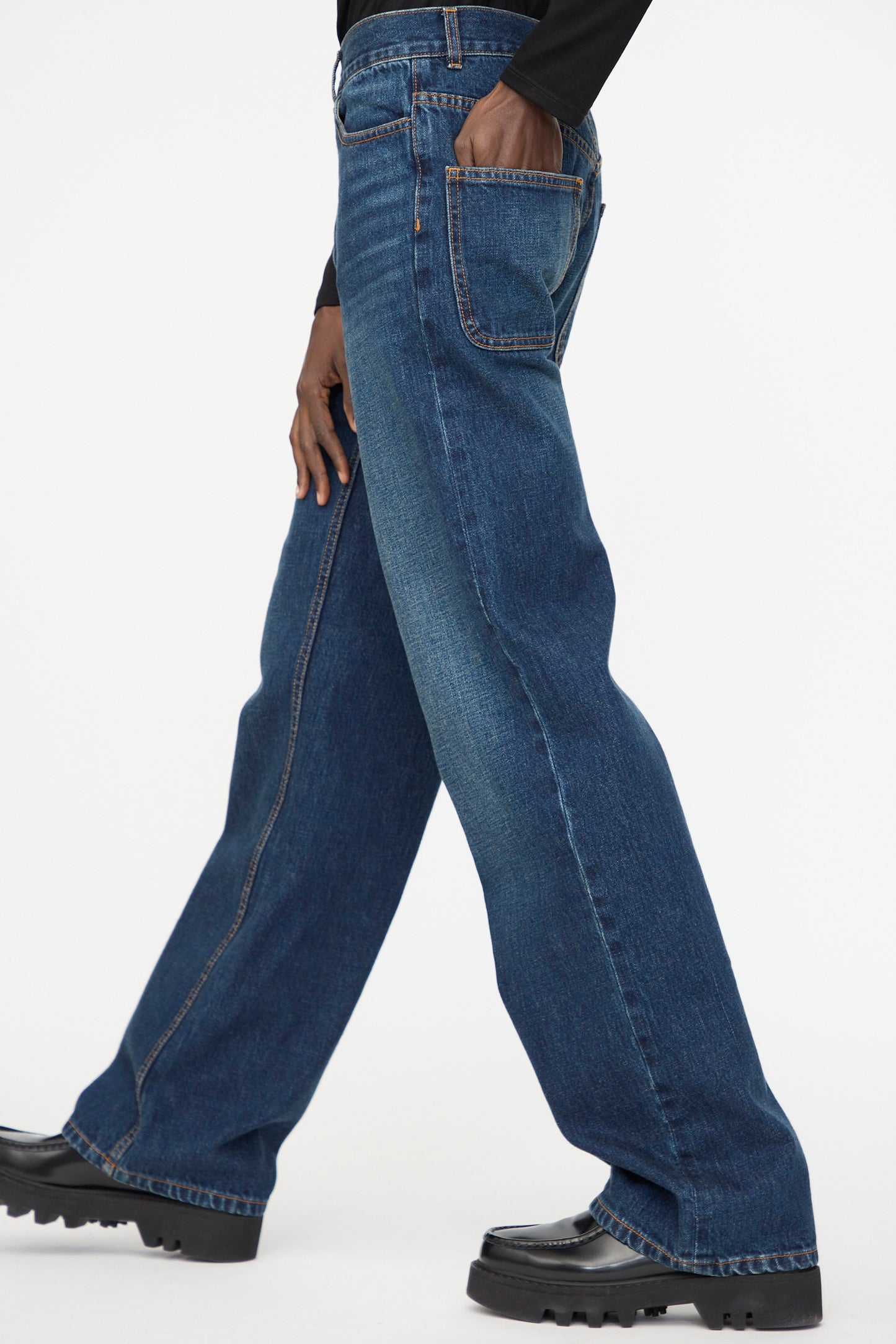 A person wearing Jesse Kamm Japanese Denim Alta Jeans in Medium Roast and black shoes, with one hand in the back pocket, walks against a plain white background.