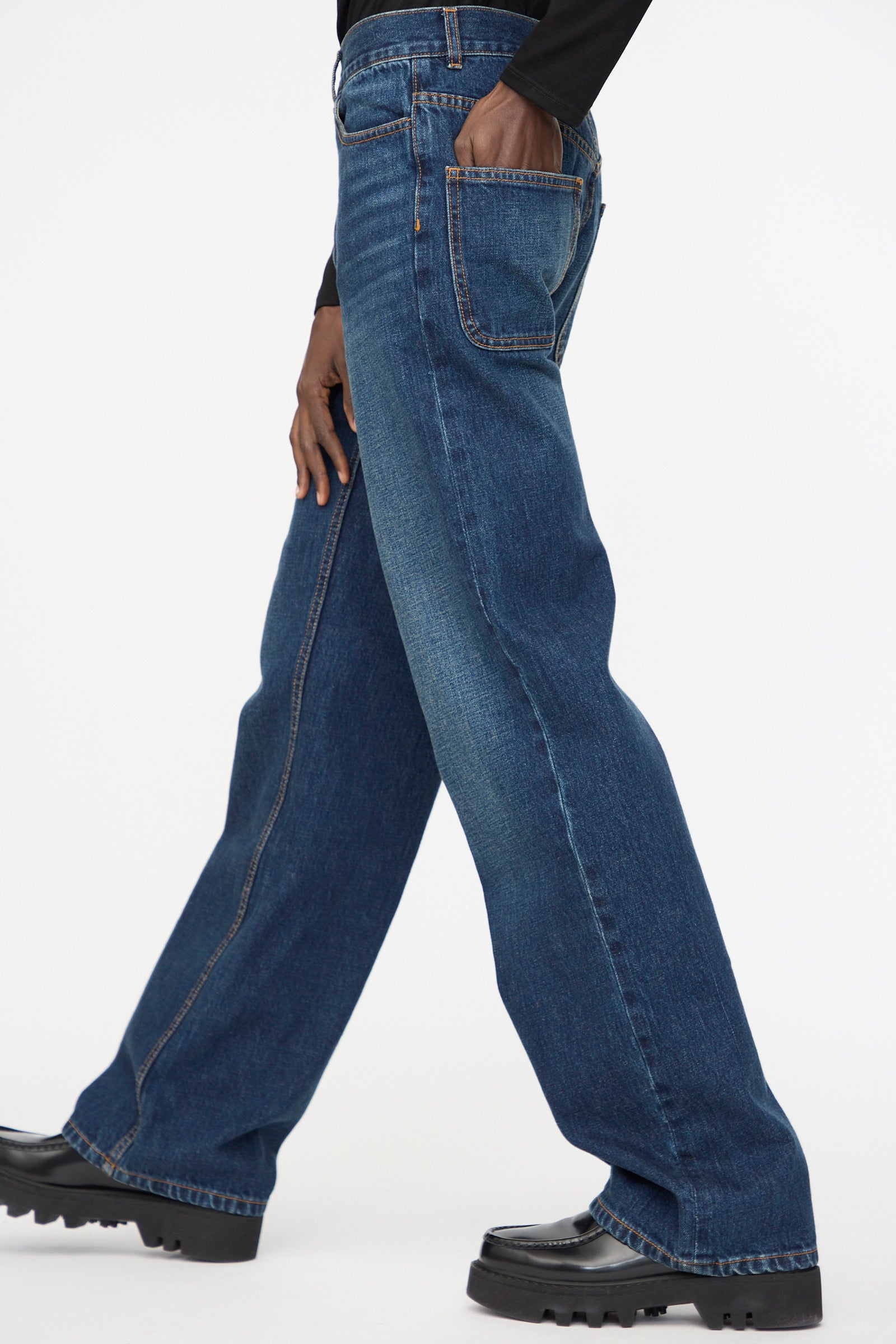 A person wearing Jesse Kamm Japanese Denim Alta Jeans in Medium Roast and black shoes, with one hand in the back pocket, walks against a plain white background.