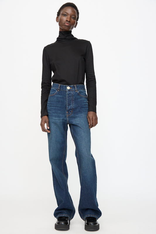 A person wears a black turtleneck, Jesse Kamm Japanese Denim Alta Jeans in Medium Roast, and black shoes while standing against a plain white background.