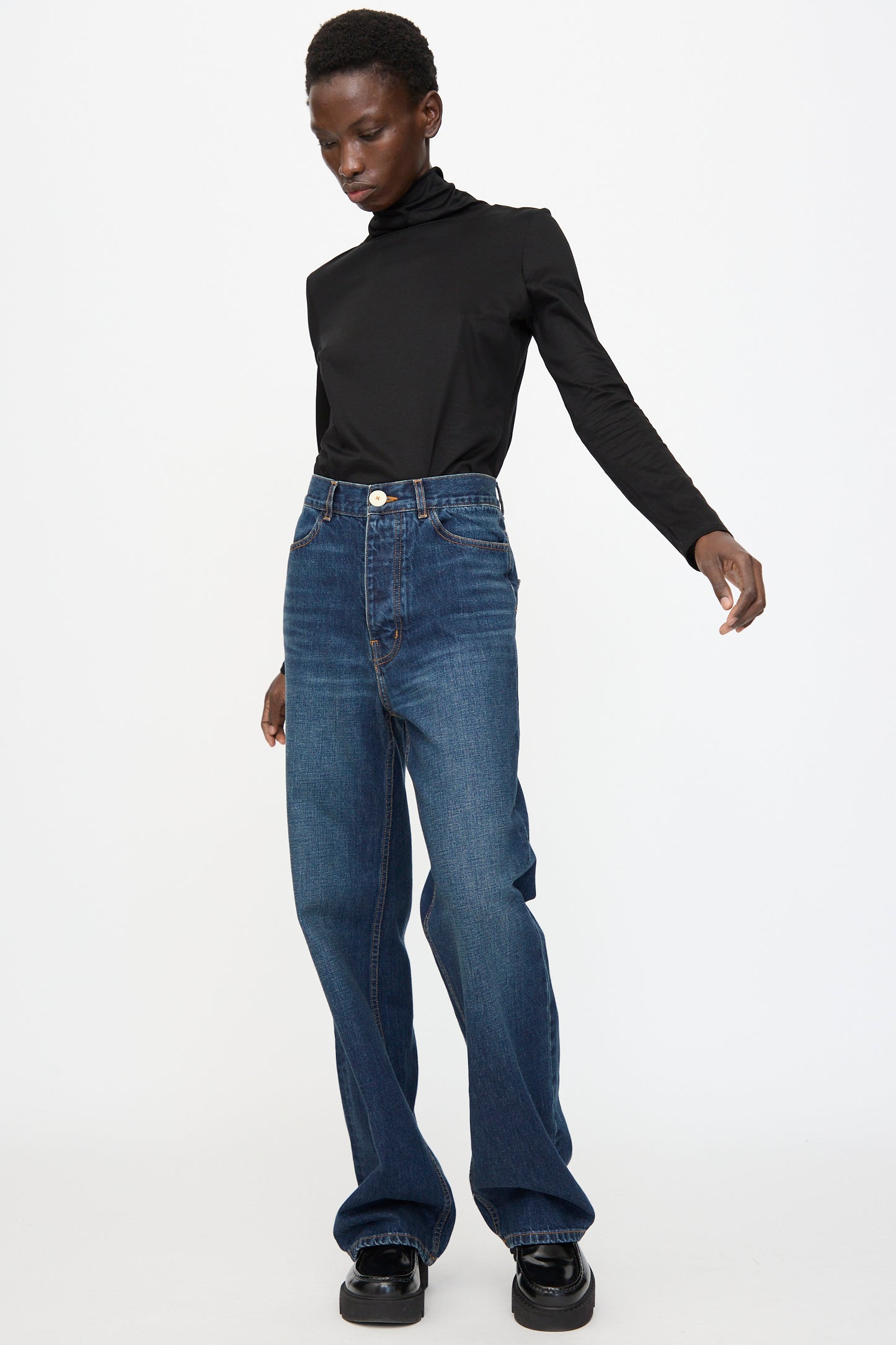 A person stands against a plain background wearing a black turtleneck, black shoes, and Jesse Kamm Japanese Denim Alta Jeans in Medium Roast, crafted from regenerative Japanese denim.