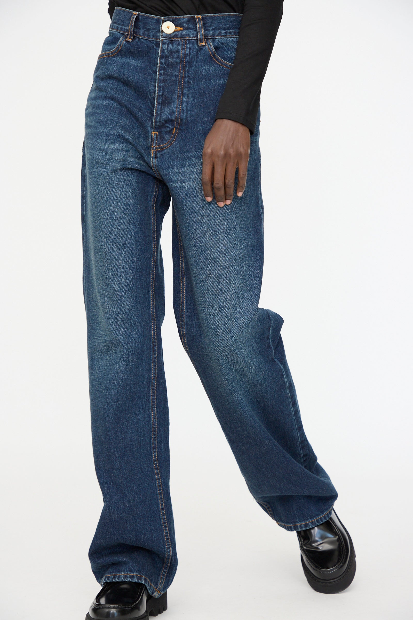 A person stands against a light background wearing Jesse Kamm’s Japanese Denim Alta Jeans in Medium Roast, featuring a high-waisted wide-leg fit, paired with a black long-sleeve top and black shoes.