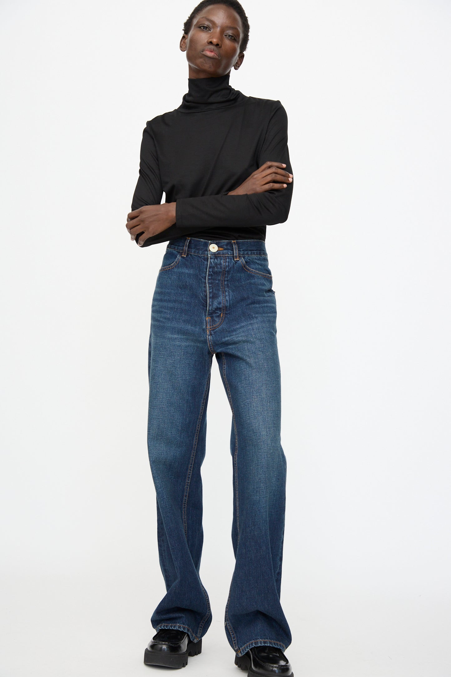 A person stands against a plain background with arms crossed, wearing a black turtleneck, black shoes, and Jesse Kamm Japanese Denim Alta Jeans in Medium Roast.