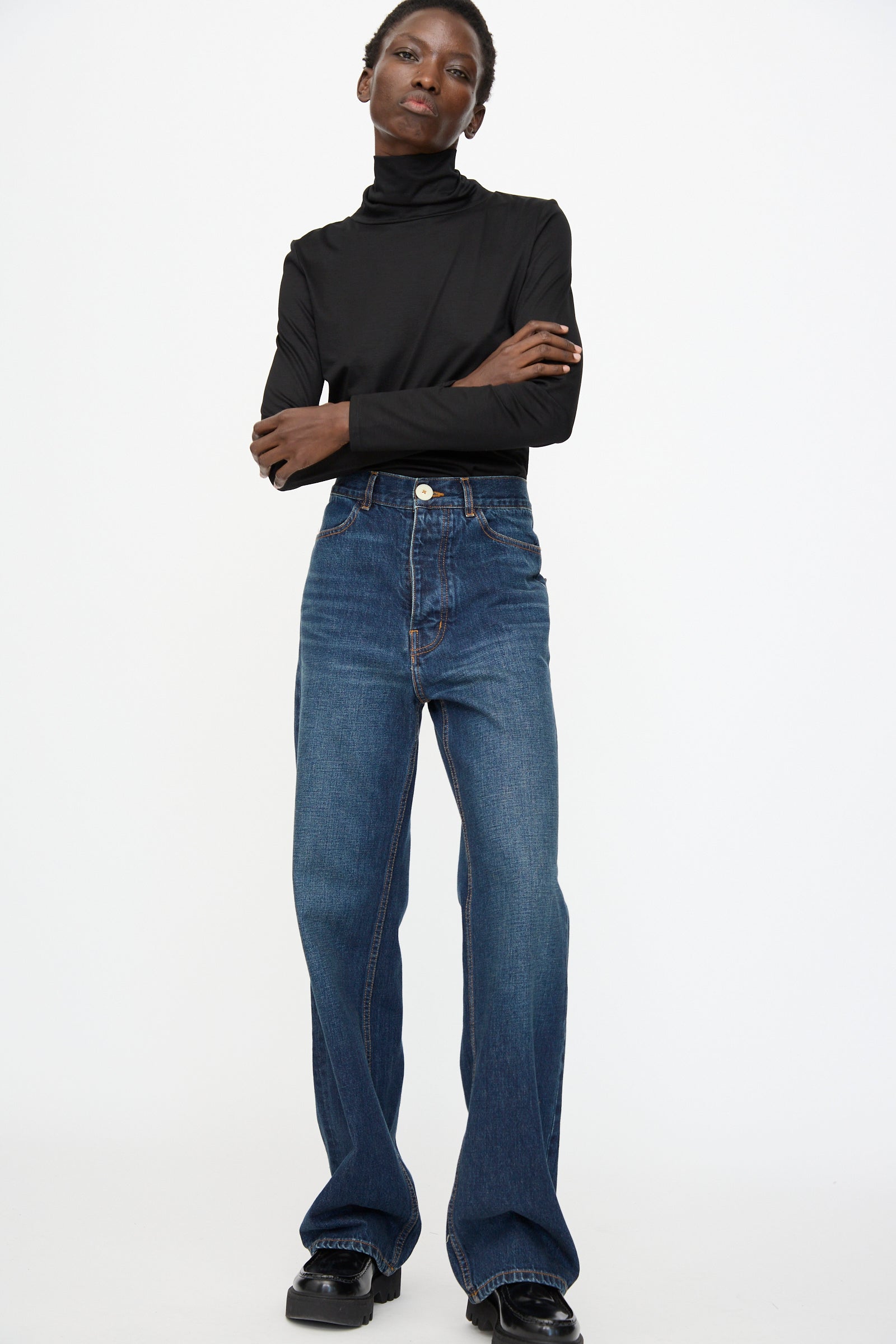 A person stands against a plain background with arms crossed, wearing a black turtleneck, black shoes, and Jesse Kamm Japanese Denim Alta Jeans in Medium Roast.