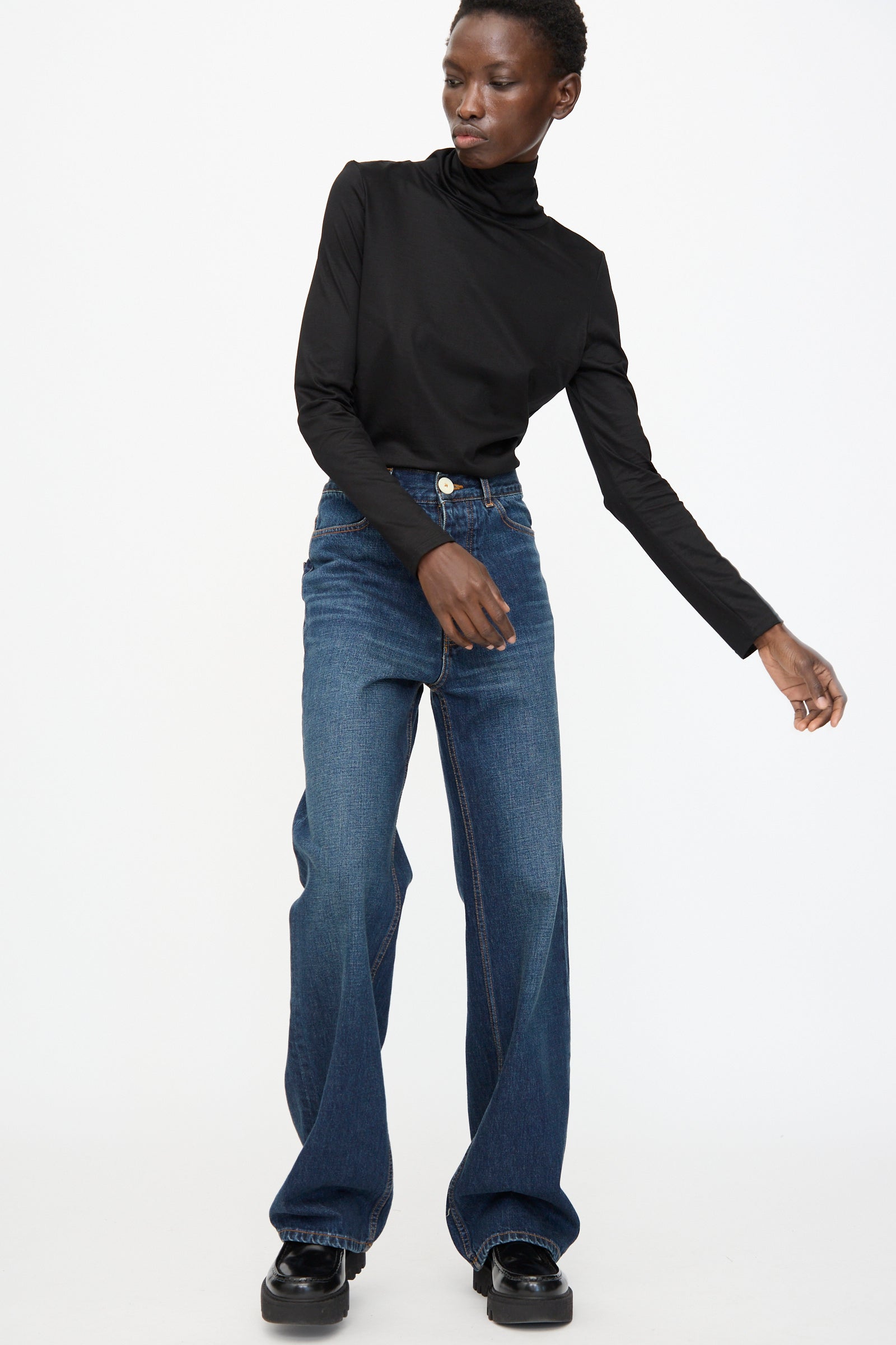 A person wearing a black turtleneck, Jesse Kamm Japanese Denim Alta Jeans in Medium Roast with a boot cut leg, and black shoes stands against a plain white background.