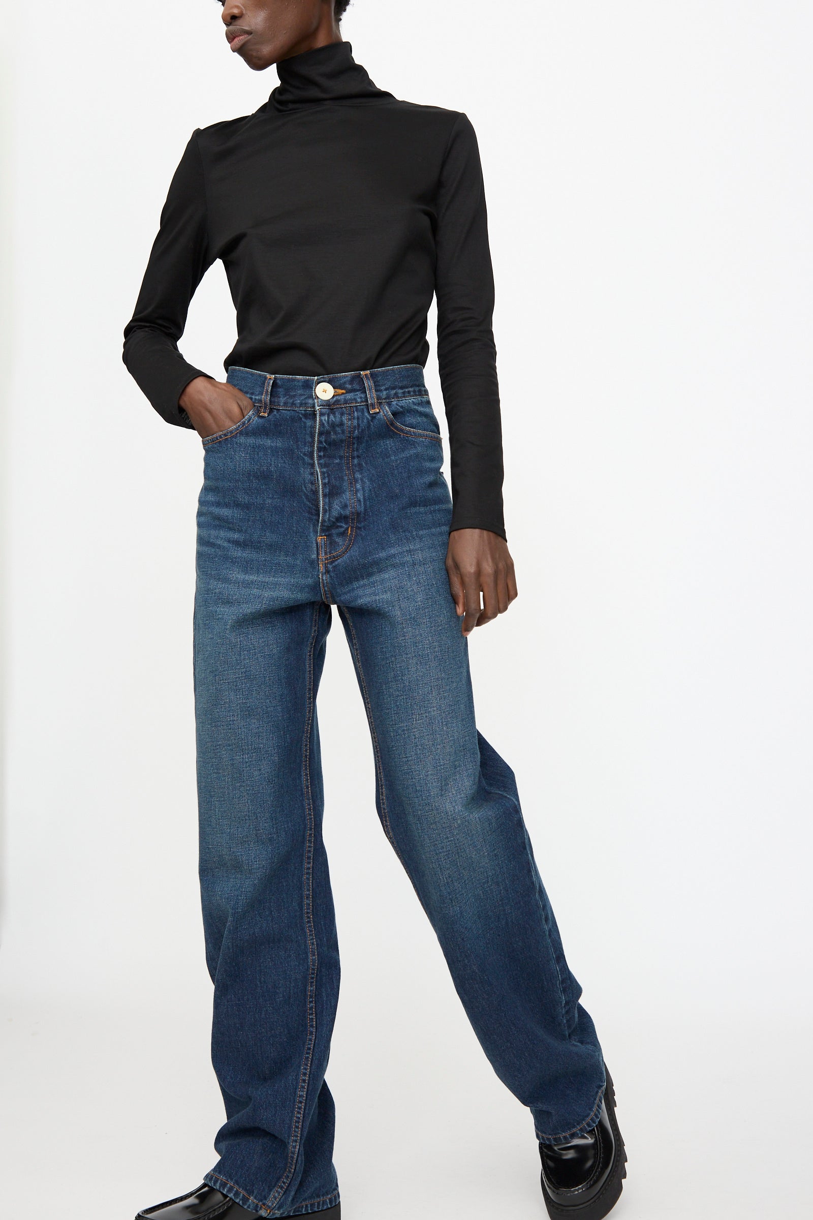 A person wears a black turtleneck long-sleeve top, Jesse Kamm Japanese Denim Alta Jeans in Medium Roast, and black shoes, standing against a plain white background.