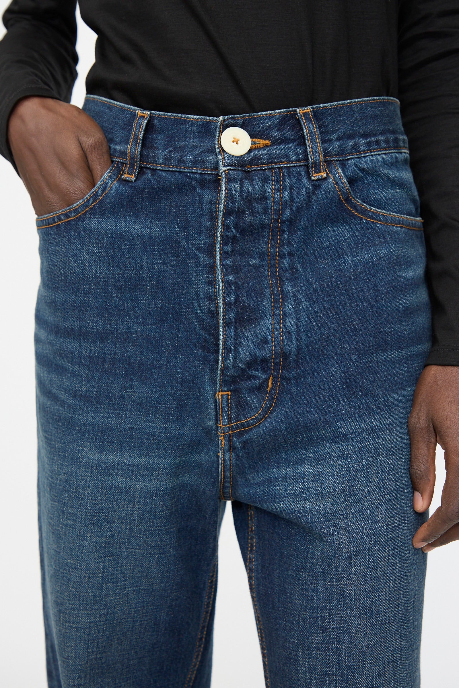 A person wearing a black long-sleeve shirt and Jesse Kamm’s Japanese Denim Alta Jeans in Medium Roast, one hand in the pocket, is shown from torso to mid-thigh.