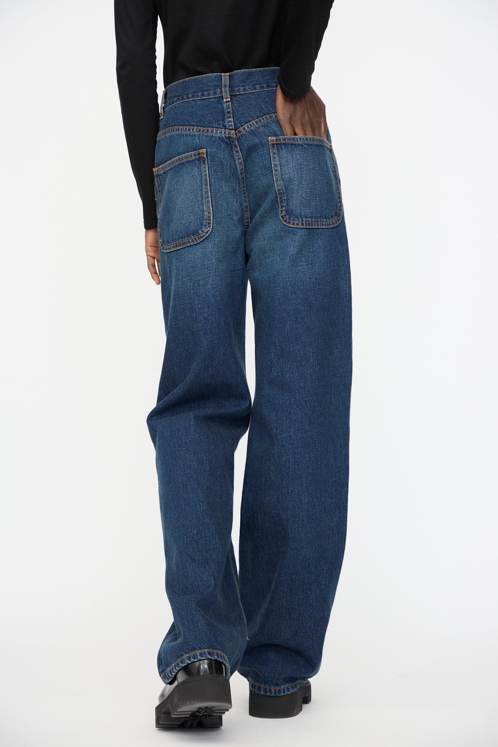 Person wearing Jesse Kamm's Japanese Denim Alta Jeans in Medium Roast—loose-fitting, high-waisted, boot cut—paired with black shoes and photographed from the back against a plain white background.
