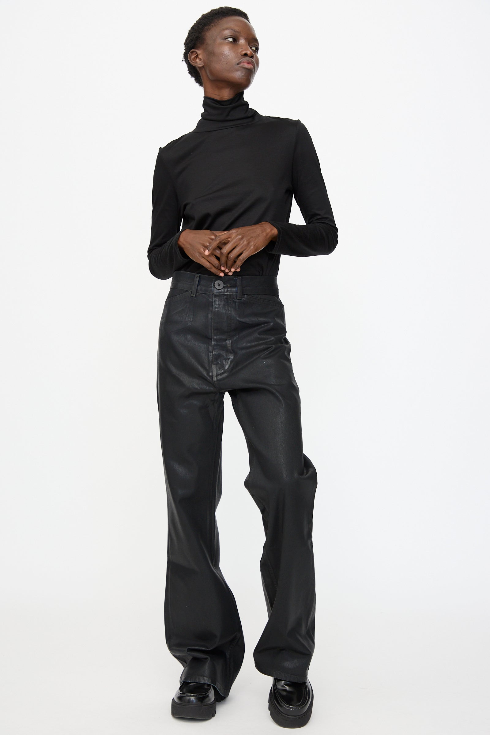 A person wearing a black turtleneck, Jesse Kamm Japanese Denim Les Deux Pants in Black Lacquer, and black platform shoes stands against a plain white background.