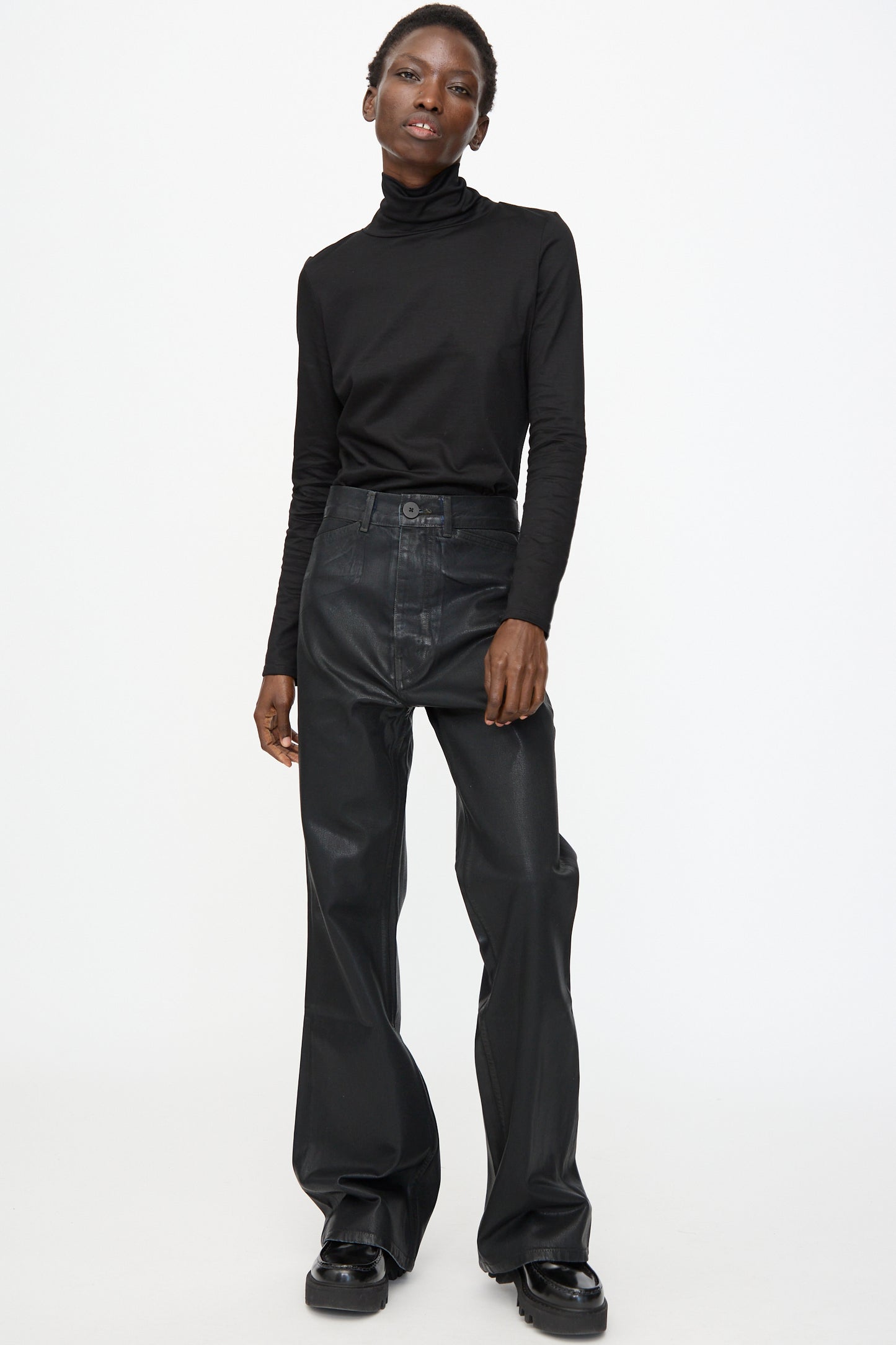 A person stands against a plain white background wearing a black turtleneck, Jesse Kamm Japanese Denim Les Deux Pants in Black Lacquer, and sleek black shoes.