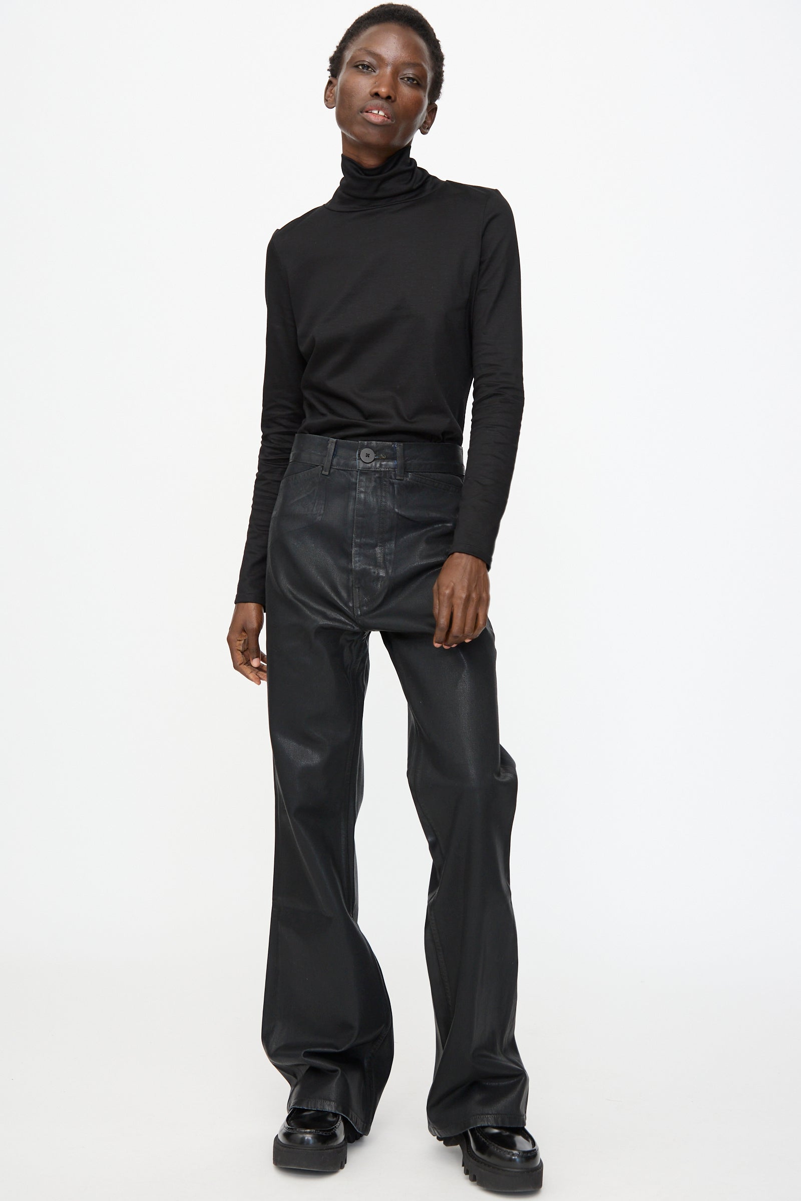 A person stands against a plain white background wearing a black turtleneck, Jesse Kamm Japanese Denim Les Deux Pants in Black Lacquer, and sleek black shoes.