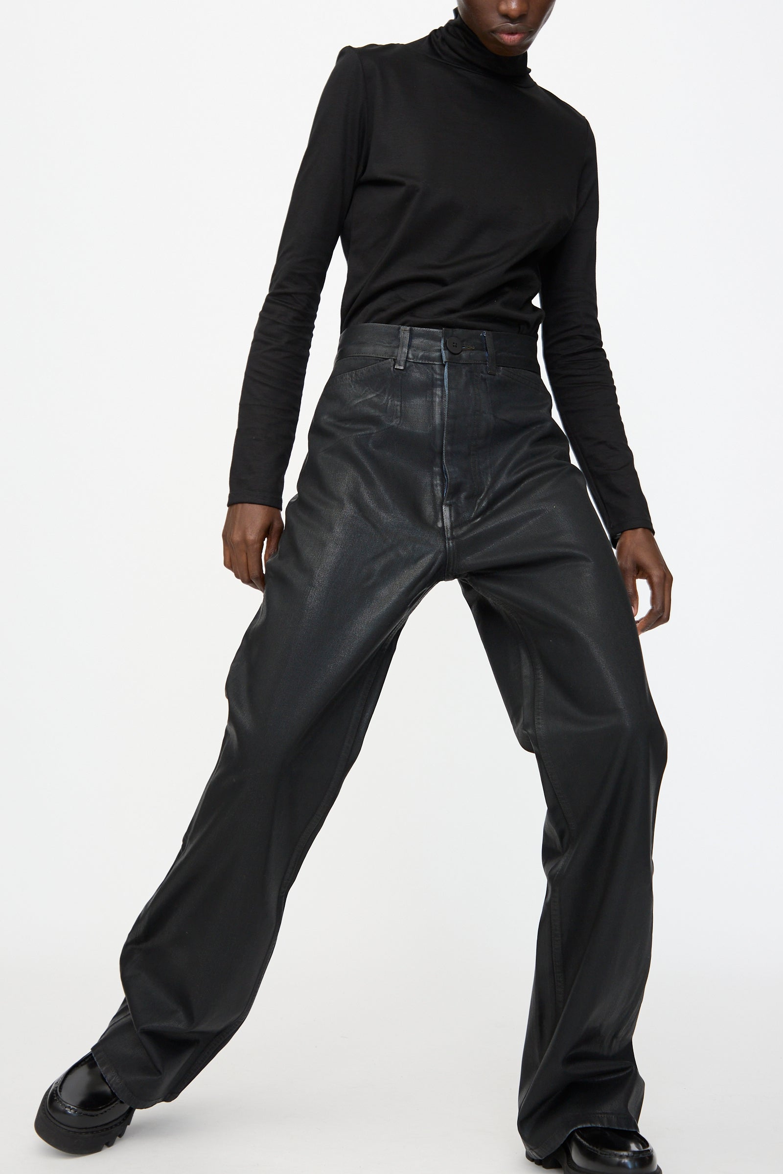 A person wearing a black turtleneck, Jesse Kamm Japanese Denim Les Deux Pants in Black Lacquer, and black shoes stands against a plain white background.