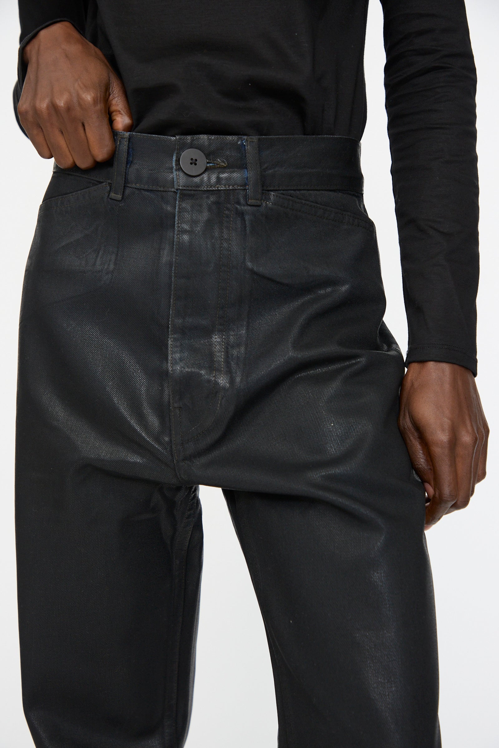 A person wearing Jesse Kamm’s Japanese Denim Les Deux Pants in Black Lacquer and a black long-sleeve shirt, with one hand on the waistband.