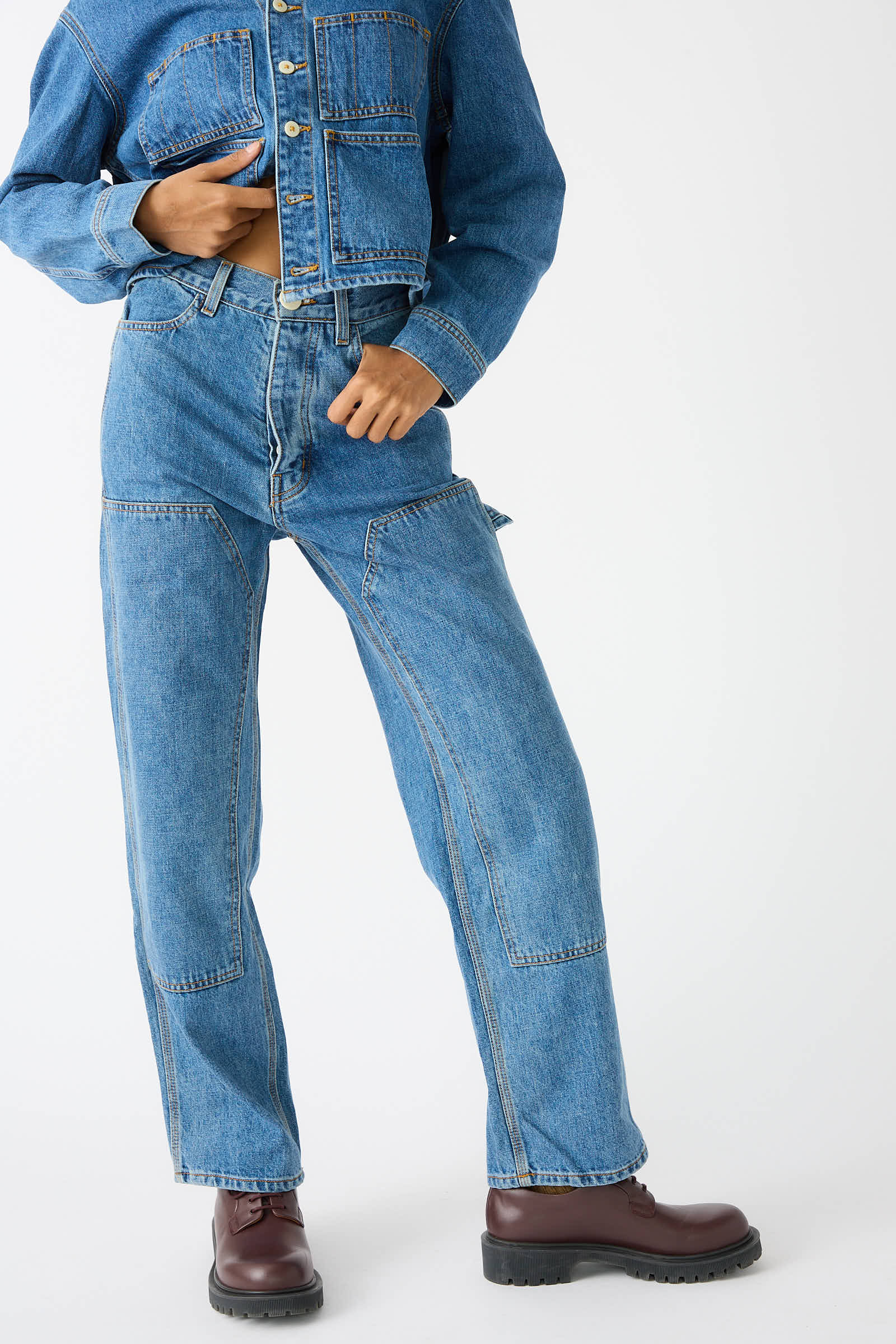 A person is wearing a denim jacket and the Japanese Denim Patchfront Handy Pant in Cowboy Blue by Jesse Kamm, paired with brown lace-up boots against a plain background.