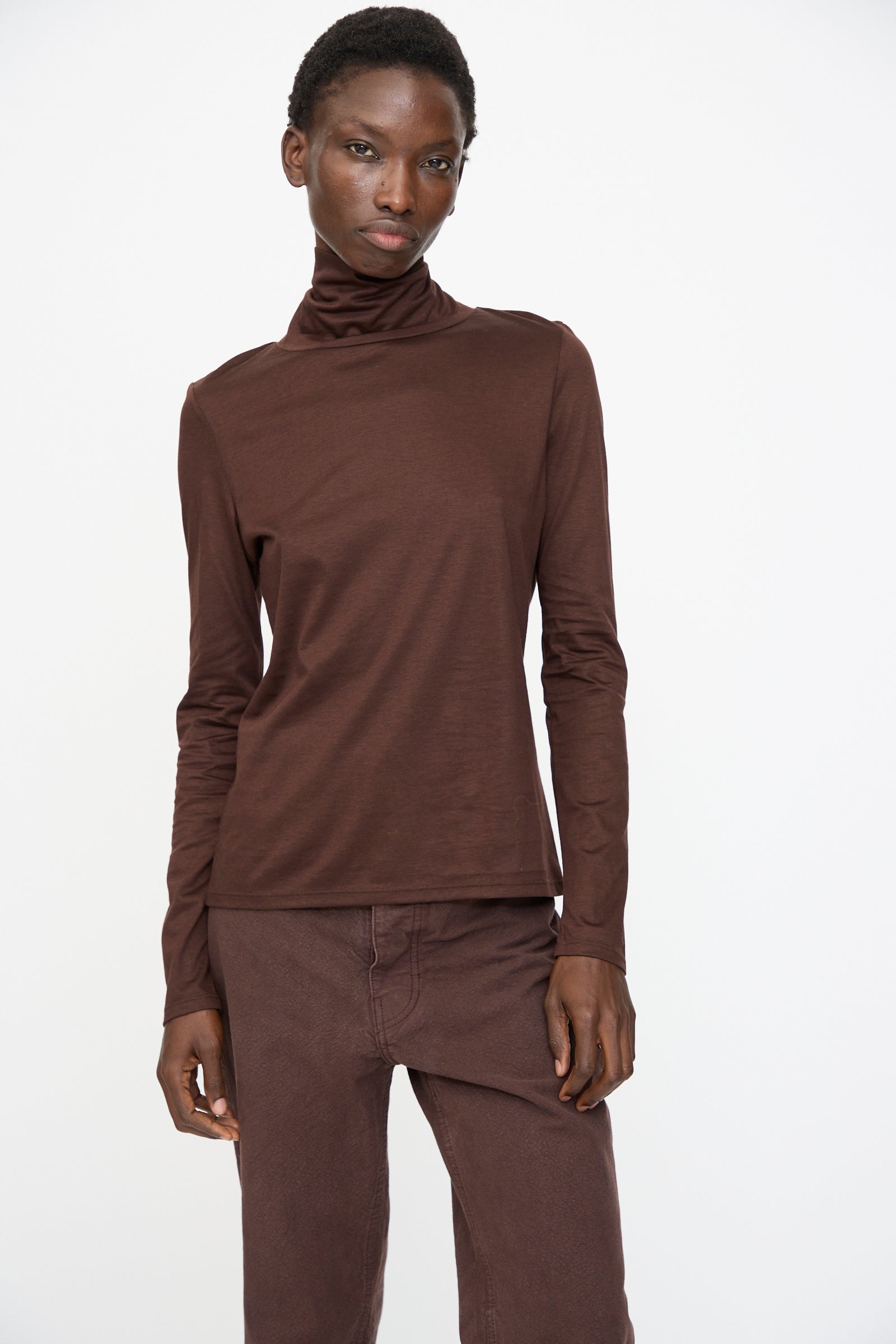 A person wears the Jesse Kamm Japanese Organic Cotton Classic Turtleneck in Raisin, paired with matching pants, both crafted from soft Japanese knit, standing against a plain white background.
