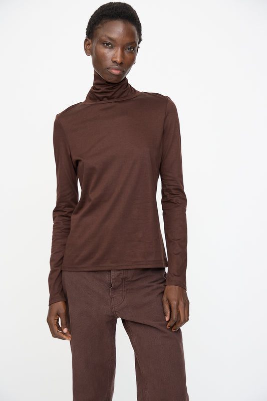A person wears the Jesse Kamm Japanese Organic Cotton Classic Turtleneck in Raisin, paired with matching pants, both crafted from soft Japanese knit, standing against a plain white background.