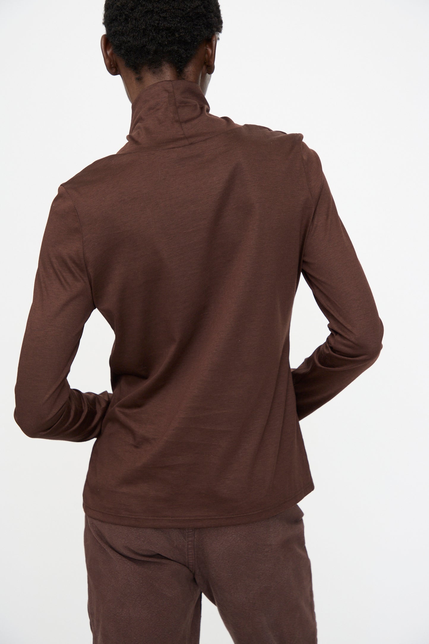 A person stands with their back to the camera, wearing the Jesse Kamm Japanese Organic Cotton Classic Turtleneck in Raisin and matching brown pants, set against a plain white background.