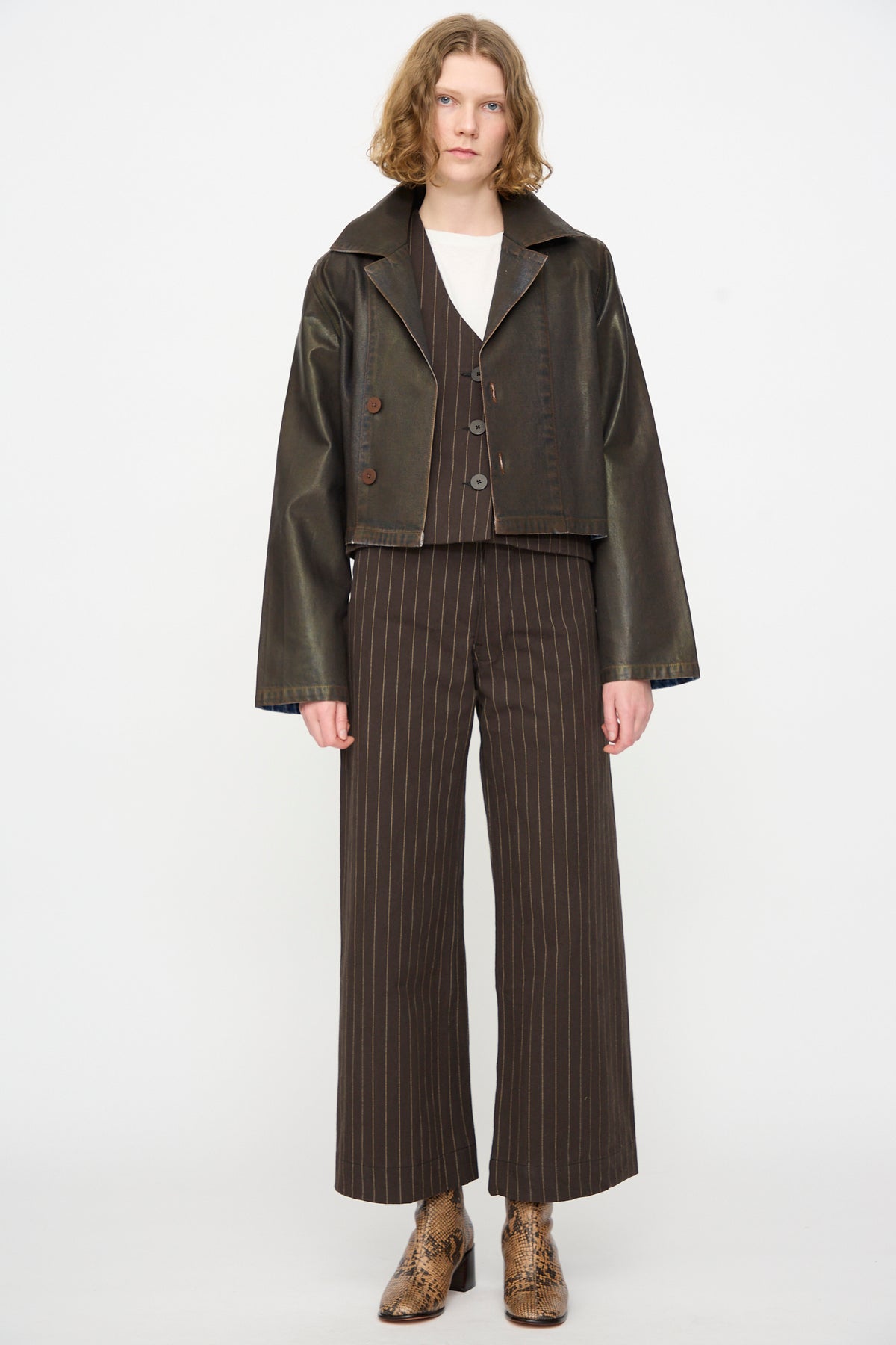 Person standing against a white background, wearing the Jesse Kamm Jones Jacket in Rich Brown Lacquer—a relaxed, dark cropped jacket—paired with a brown pinstripe suit, white top, and snakeskin boots.