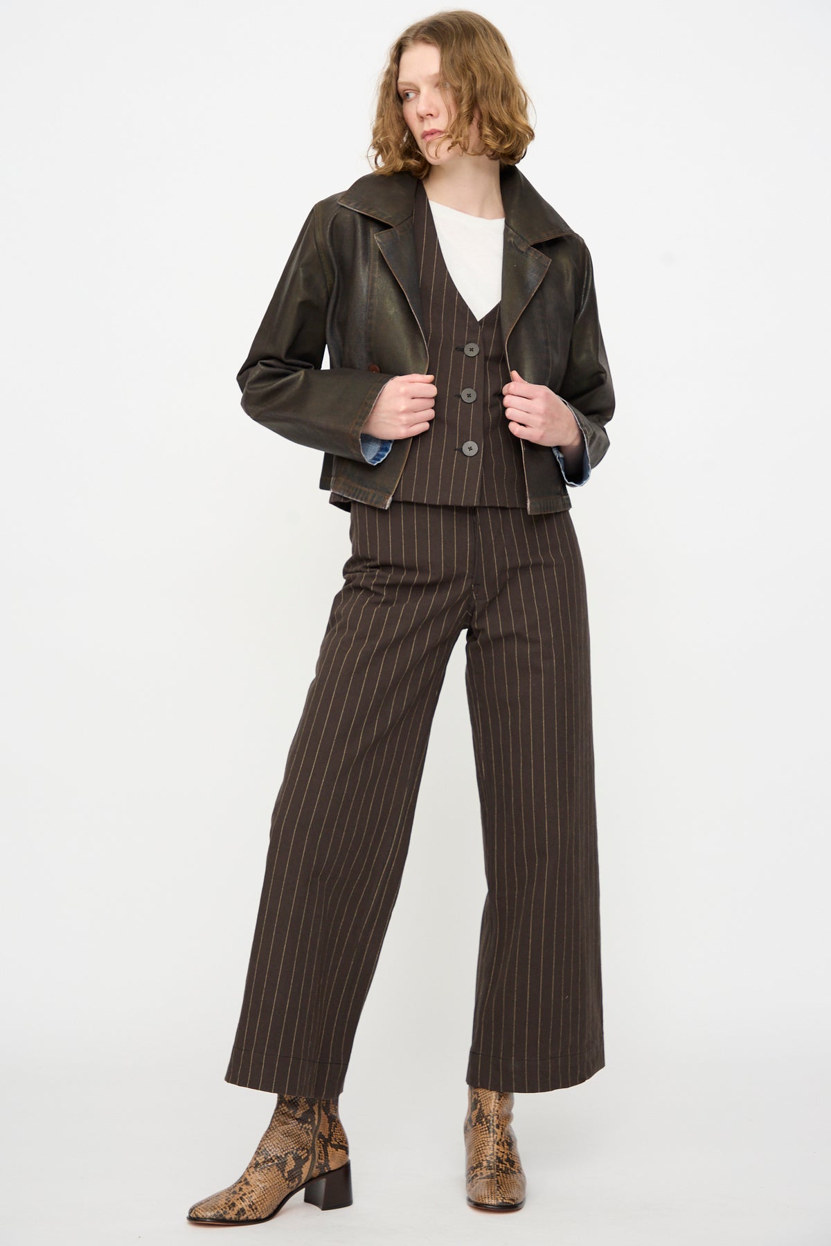 A person wearing the Jesse Kamm Jones Jacket in Rich Brown Lacquer over a pinstriped vest and pants with a white shirt and snake print boots, standing against a plain white background.