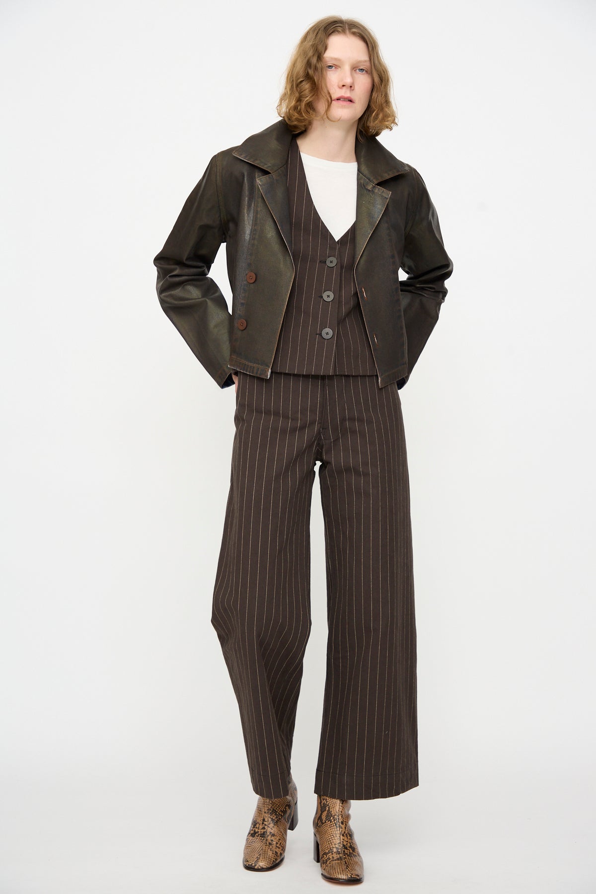 Wearing the Jones Jacket in Rich Brown Lacquer by Jesse Kamm layered over a brown pinstripe suit with a matching vest and white shirt, this person completes the look with snakeskin-patterned ankle boots, standing against a plain white background.