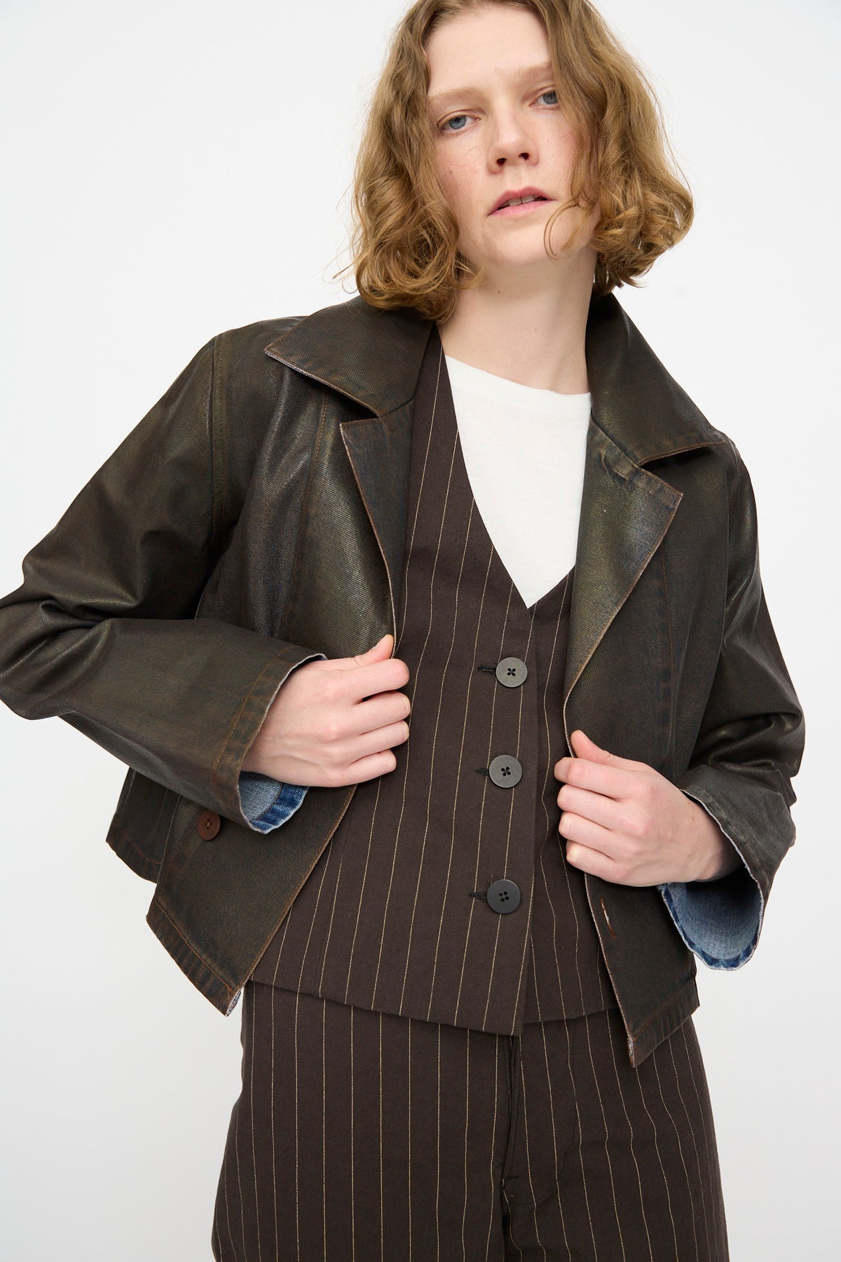 A person wearing a dark brown pinstripe suit with a white shirt and the Jesse Kamm Jones Jacket in Rich Brown Lacquer—a cropped, relaxed piece crafted from regenerative Japanese denim—stands against a plain light background.